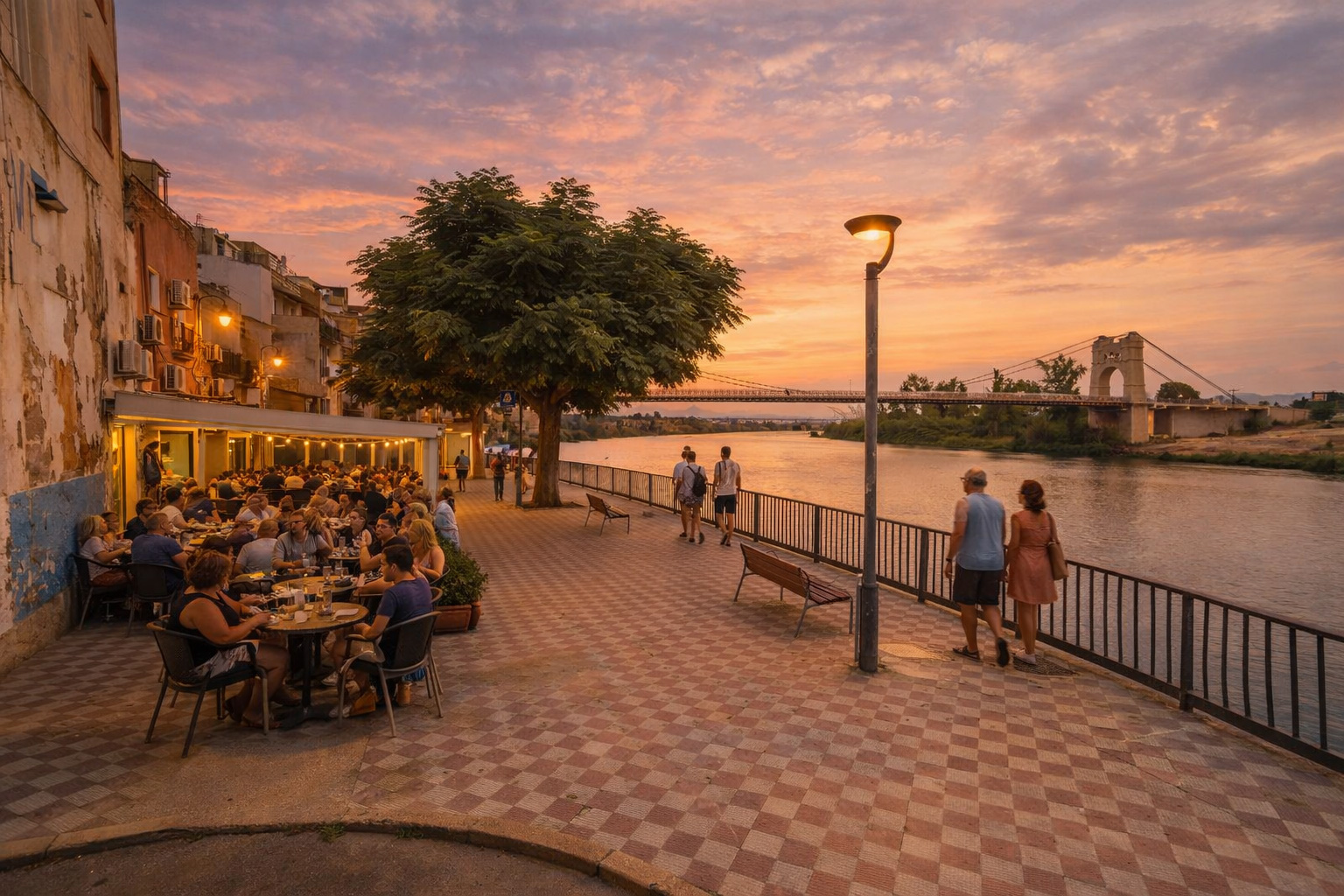 Abendstimmung am Carrer Poador in Amposta an der Uferpromenade des Ebro, mit gemütlichem Straßencafé links an der Mauer voller Gäste, eingeschalteten Laternen und warm beleuchteten Fenstern, während mehrere Spaziergänger am Geländer entlanggehen und der Fluss im Licht des Sonnenuntergangs bis zur Brücke im Hintergrund schimmert.