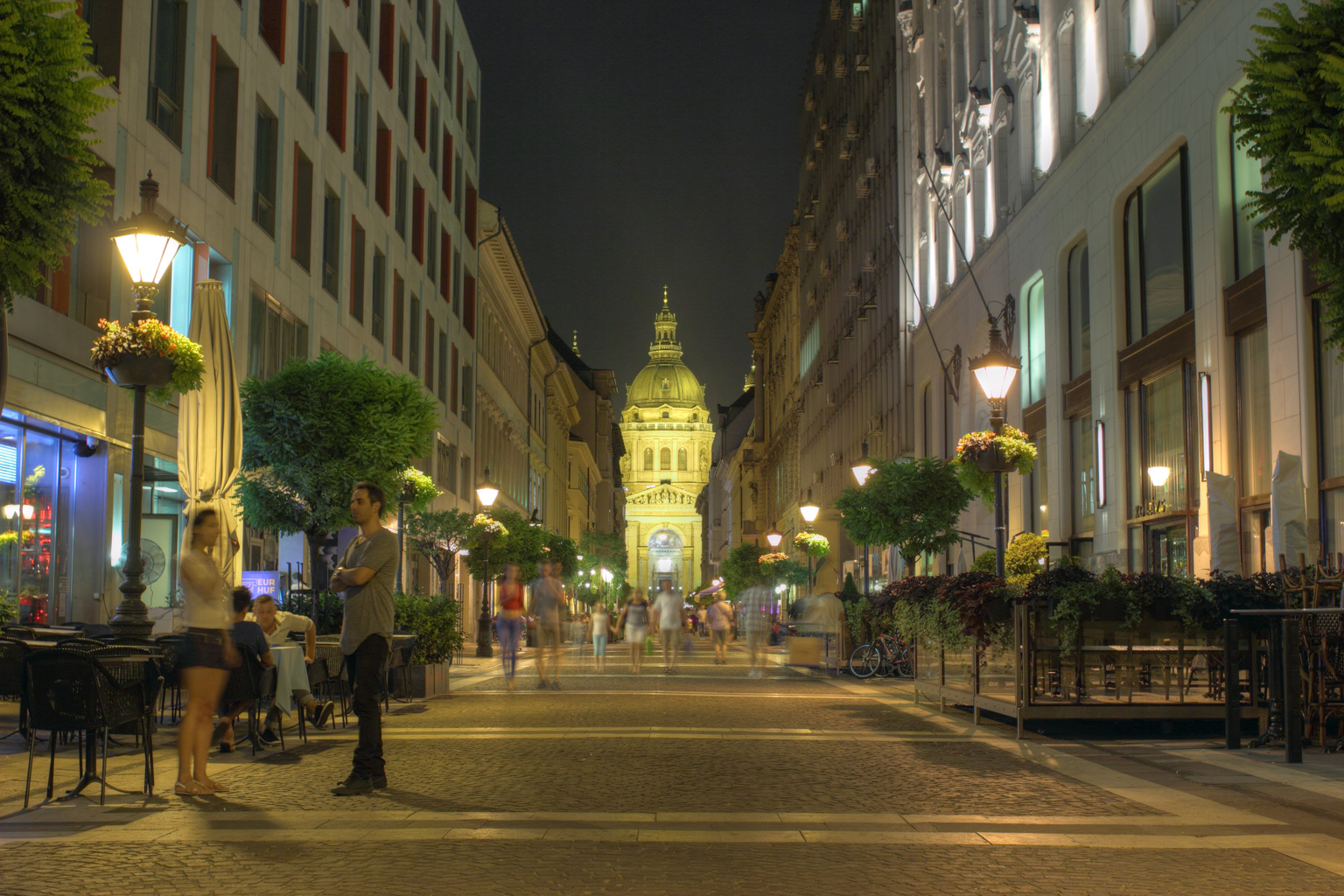 Abendstimmung in der Altstadt von Budapest mit beleuchtetem Straßencafé, hell erleuchteten Schaufenstern, Straßenbeleuchtung und Spaziergängern vor malerischem Sonnenuntergangshimmel.