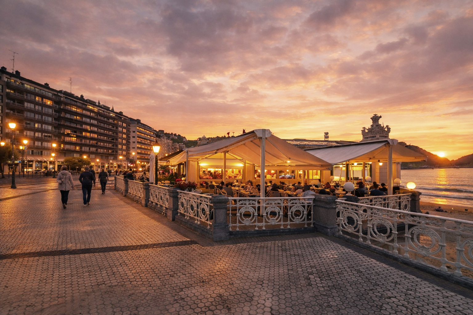 Abendstimmung an der Kontxa Pasealekua mit beleuchtetem Strandcafé unter weißen Schirmen, Spaziergängern auf der Promenade, eingeschalteter Straßenbeleuchtung und Blick auf die Bucht von La Concha bei Sonnenuntergang