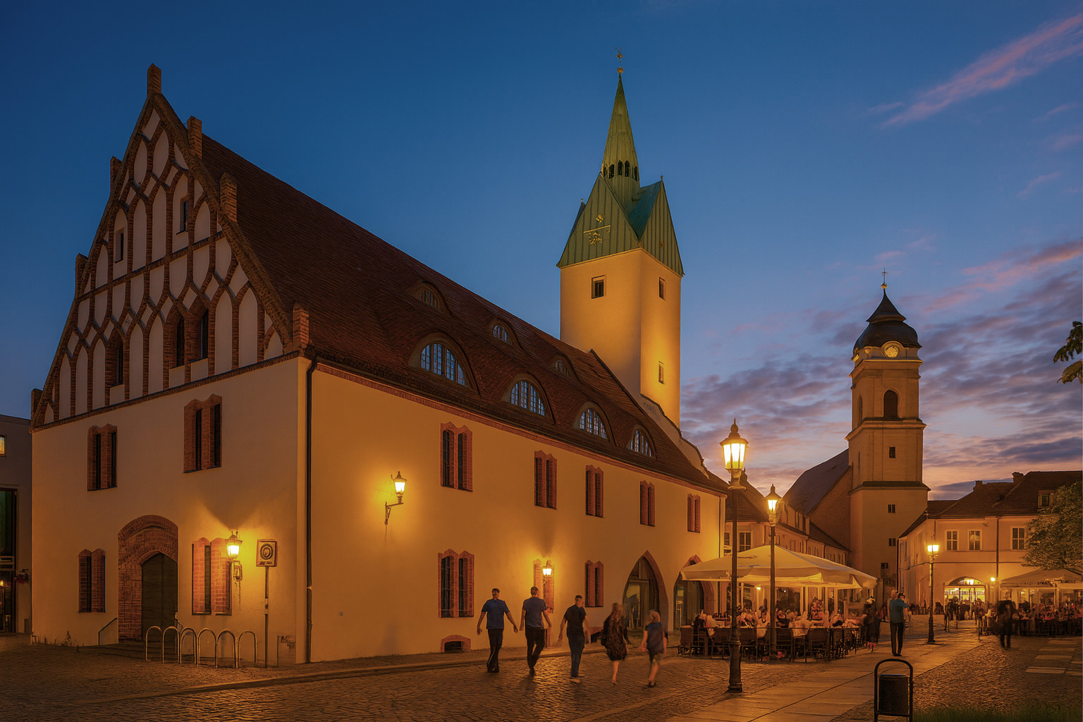 Abendstimmung am Rathaus in Fürstenwalde mit beleuchteter Fassade, Straßencafé und Spaziergängern.