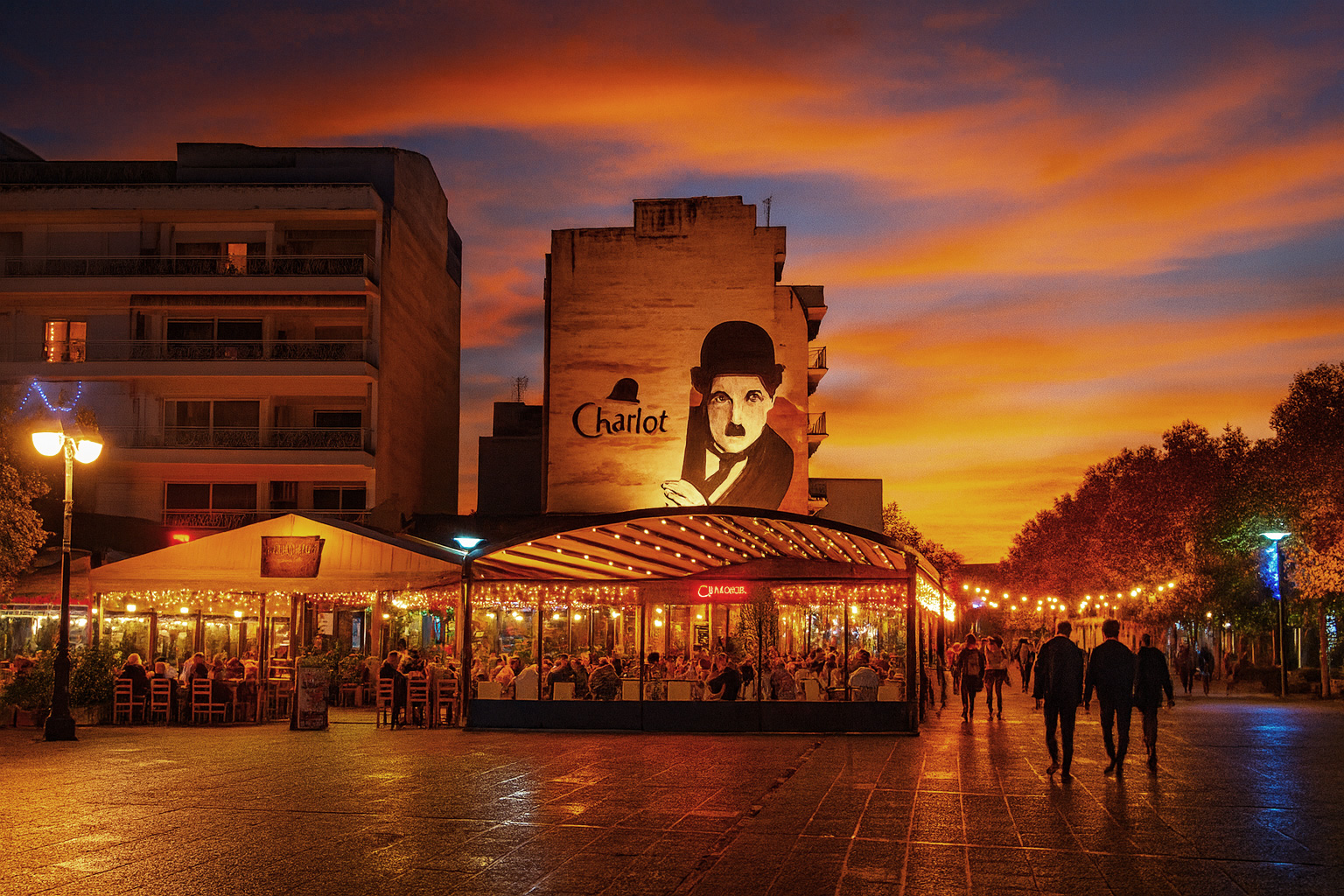 Dimitri Tertipi Platz in Karditsa bei Sonnenuntergang mit beleuchtetem Charlot-Café, Spaziergängern und warmem Abendlicht.