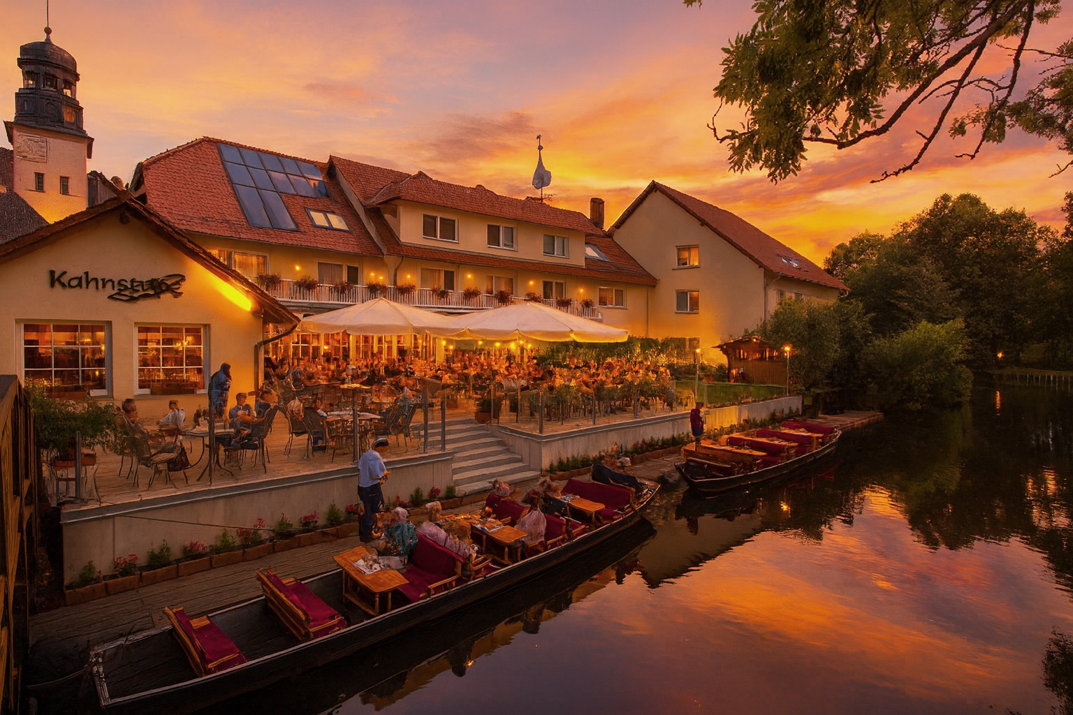 Früher Abend am Spreeufer in Lübben mit gut besuchtem Ufercafé; leere Kähne am Wasser, warmes Licht und malerischer Himmel.