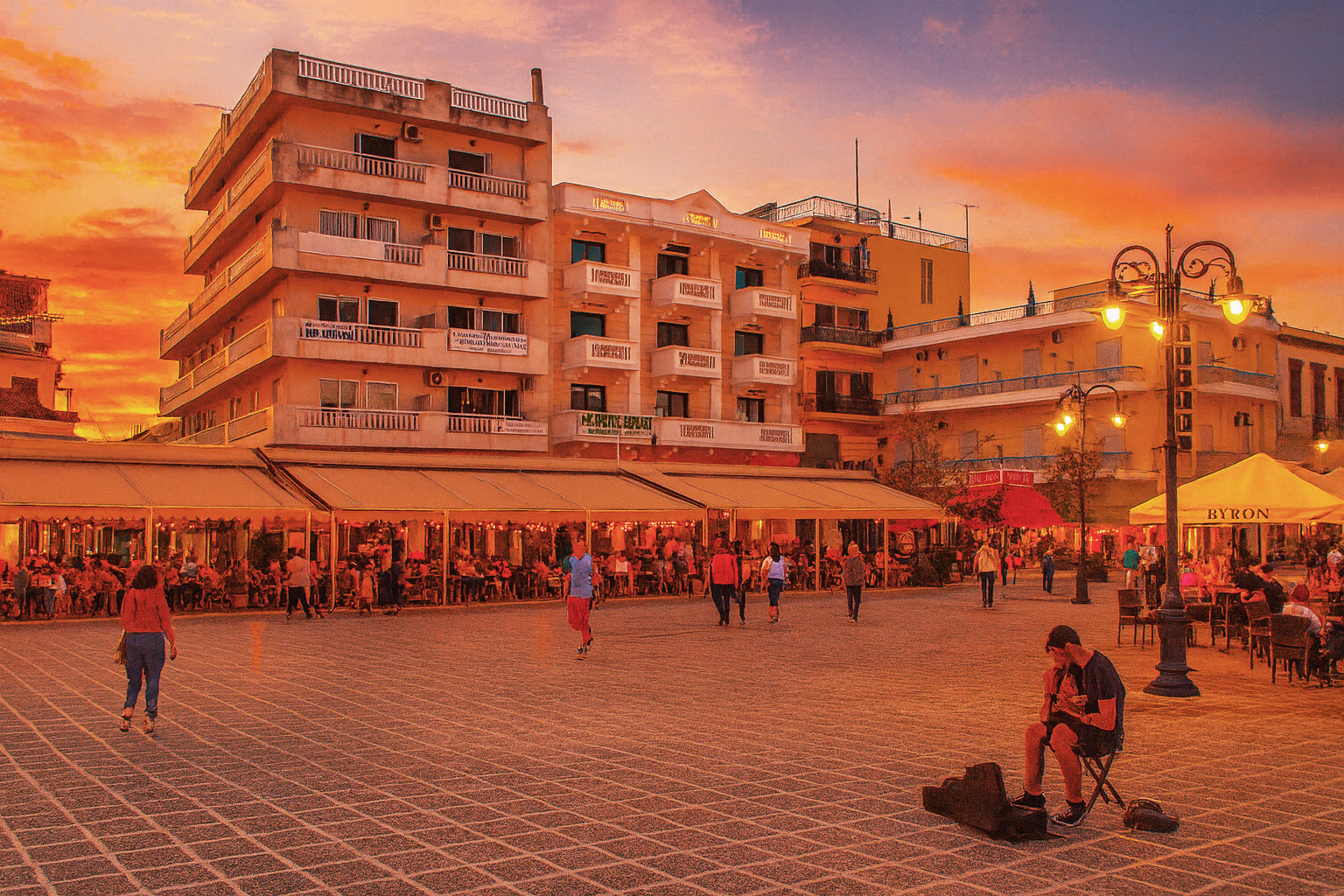 Abendstimmung auf dem Stadtplatz von Mesolongi mit beleuchtetem Straßencafé, Spaziergängern und einem Gitarristen als Straßenmusiker unter Laternen.