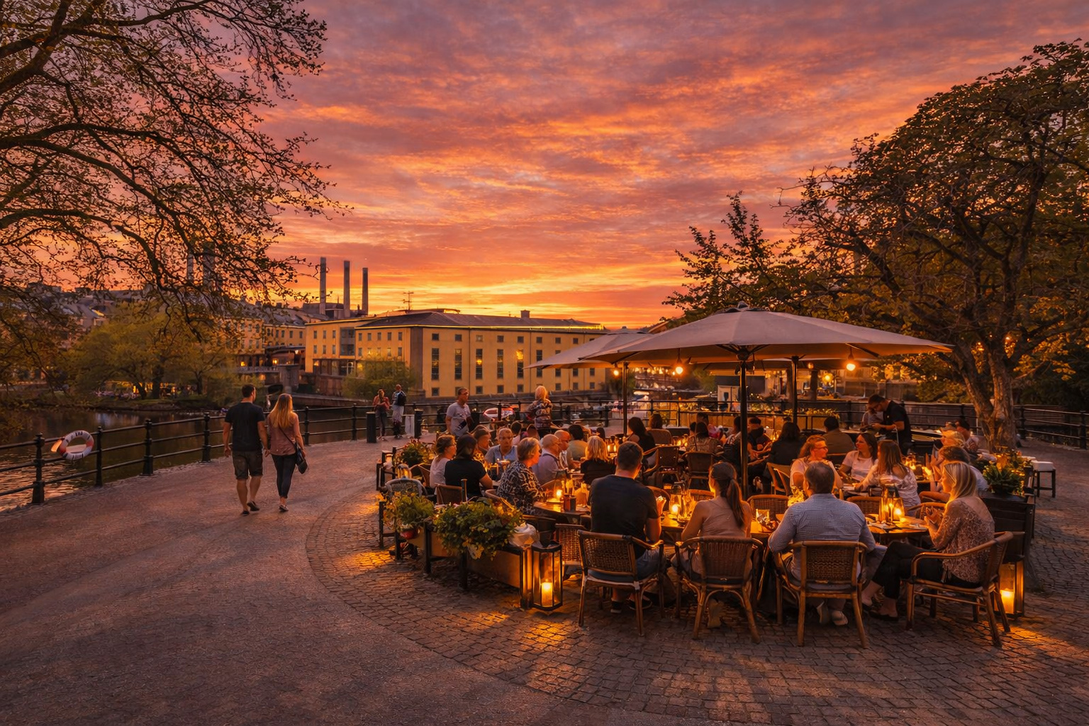 Järnbrogatan in Norrköping bei Sonnenuntergang mit Blick auf den Fluss und gelbe Industriegebäude im Hintergrund, warm beleuchteter Uferpromenade und eingeschalteten Laternen, belebtem Straßencafé an der gebogenen Sitzbank mit vielen Gästen unter großen Sonnenschirmen sowie Spaziergängern entlang des Weges, Bäume rahmen die Szene vor einem orangefarbenen Abendhimmel