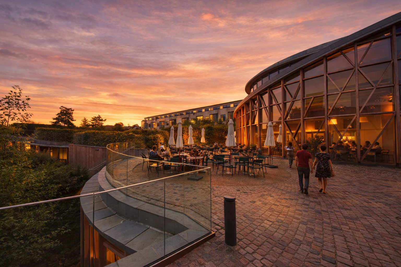 H.C. Andersen Haven in Odense bei Sonnenuntergang mit moderner Glasfassade und Holzstreben, warm beleuchtetem Innenraum, eingeschalteter Außenbeleuchtung und belebter Caféterrasse mit Tischen, Stühlen und weißen Sonnenschirmen, Spaziergänger auf dem gepflasterten Weg entlang der geschwungenen Glasbrüstung