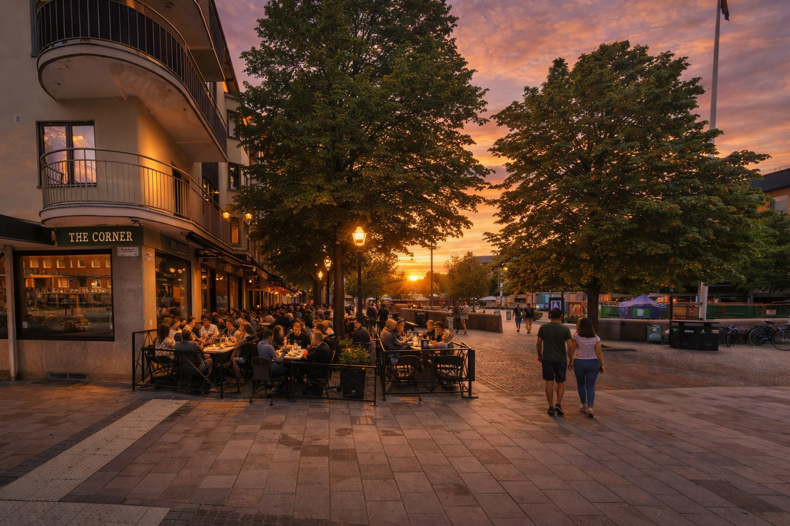 Belebtes Straßencafé an der Kanalgatan in Skellefteå bei Sonnenuntergang mit warm erleuchteten Fenstern, voll besetzten Außentischen unter Bäumen, eingeschalteter Straßenlaterne und Spaziergängern auf dem gepflasterten Platz vor modernen Gebäuden