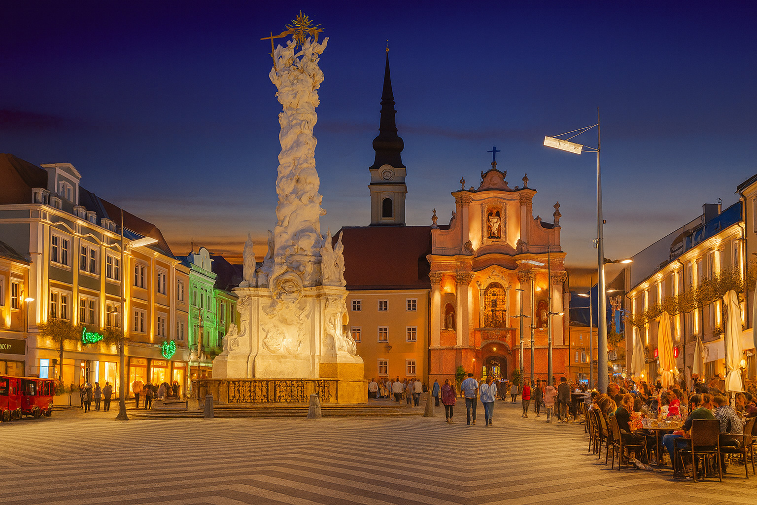Abendstimmung am Hauptplatz in St. Pölten mit der Dreifaltigkeitssäule im Vordergrund, beleuchtetem Straßencafé, Spaziergängern und farbenfrohem Sonnenuntergangshimmel.
