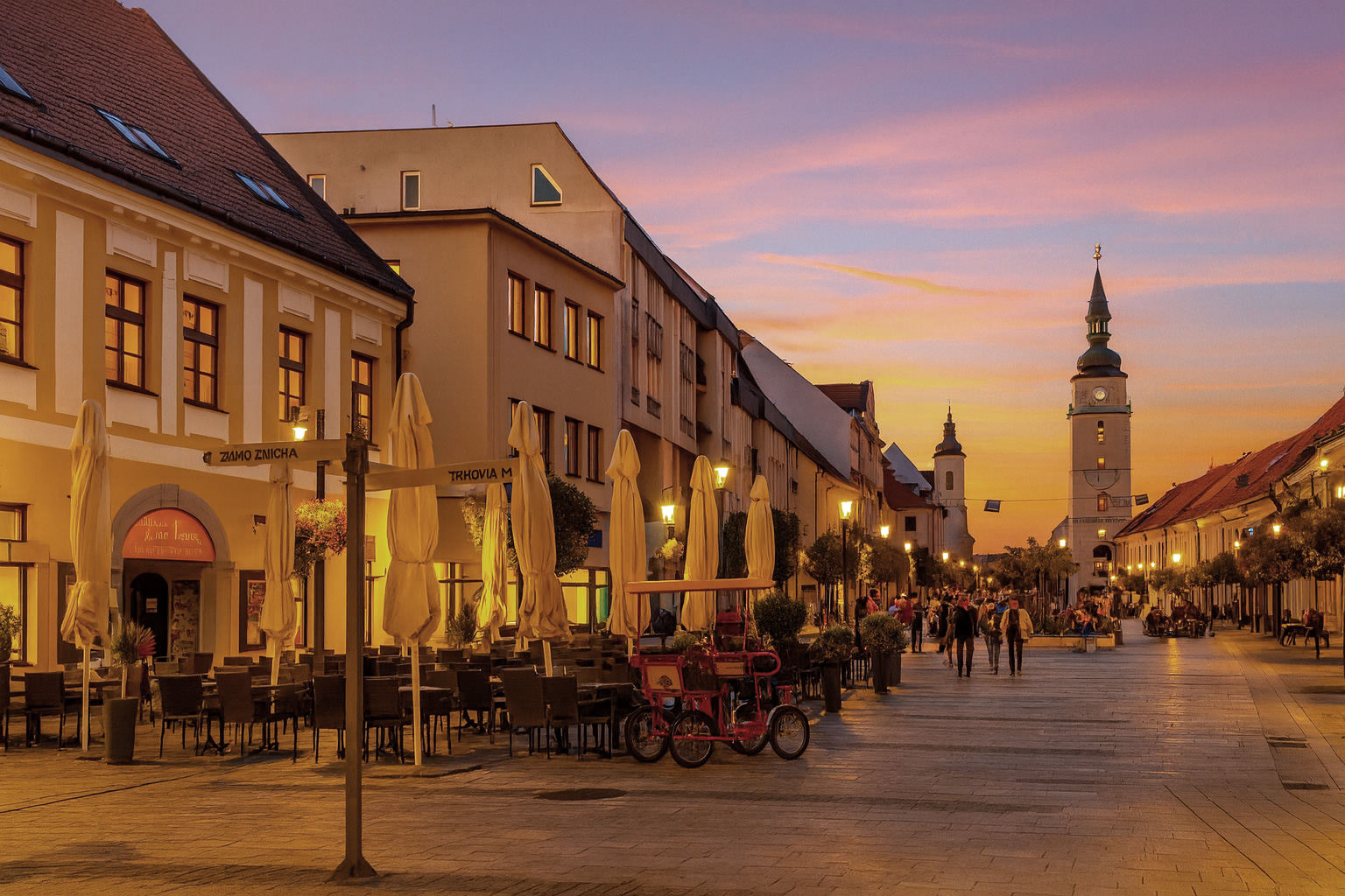 Abendstimmung in der Altstadt von Trnava mit hell erleuchteten Schaufenstern, belebtem Straßencafé, Spaziergängern und farbenprächtigem Sonnenuntergangshimmel über dem Uhrturm.