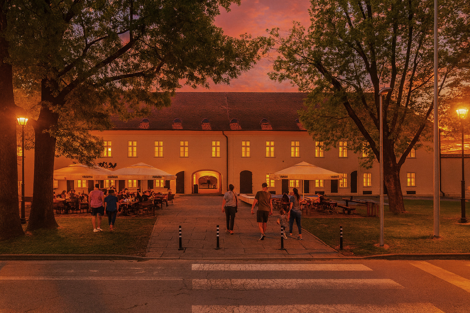 Abendstimmung am Schlosspark von Virovitica mit beleuchtetem Straßencafé unter Sonnenschirmen, Spaziergängern, erleuchteten Fenstern und malerischem Sonnenuntergangshimmel.