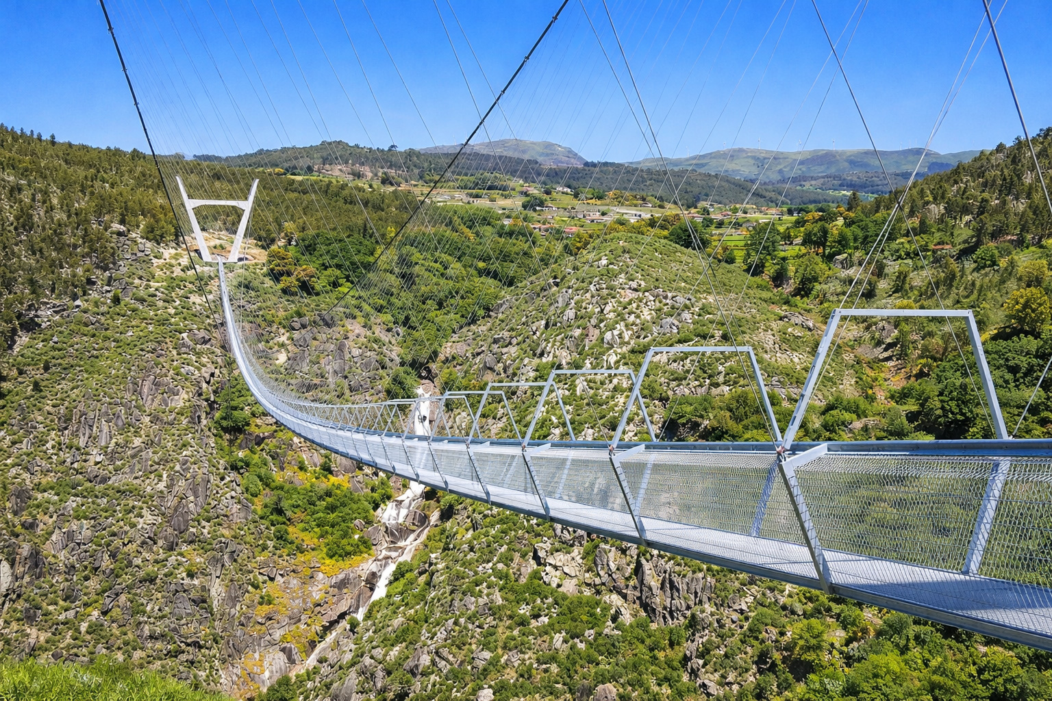 Blick entlang der Hängebrücke 516 Arouca mit Stahlseilen und Gittergeländern über einer tiefen Schlucht, darunter ein Flussbett und felsige Hänge, im Hintergrund grüne Hügel, Wälder und ein kleines Dorf unter klarem blauem Himmel