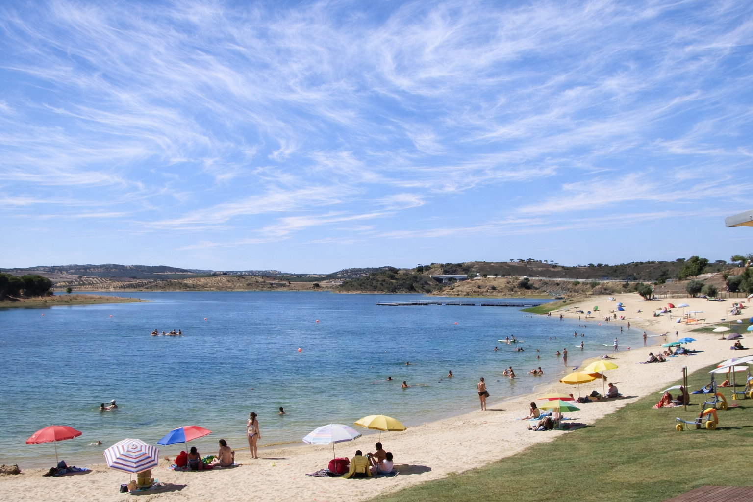 Sandstrand an der Albufeira do Azibo mit Badenden im klaren Wasser, farbigen Sonnenschirmen und Liegeflächen am Ufer, dahinter sanfte Hügel und ein Steg auf dem See unter einem Himmel mit feinen, schleierartigen Zirruswolken