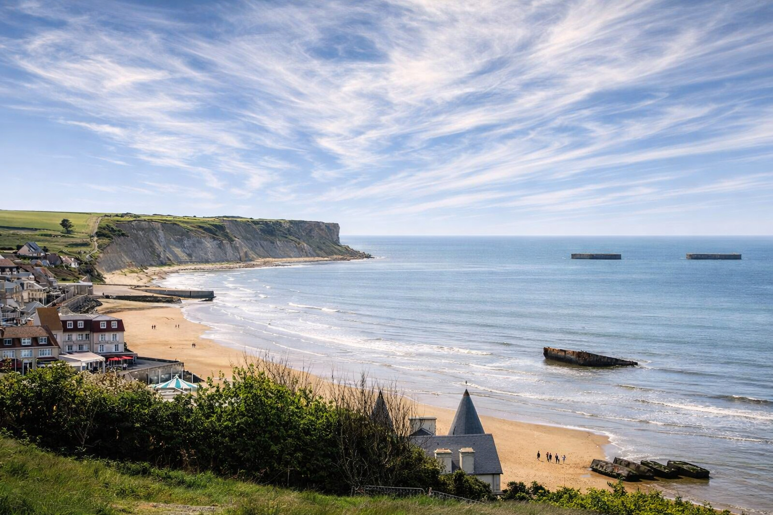 Blick über den Strand von Arromanches-les-Bains mit Küstenort, steilen Kreidefelsen und den Überresten des Mulberry-Hafens im ruhigen Ärmelkanal unter hellem Himmel