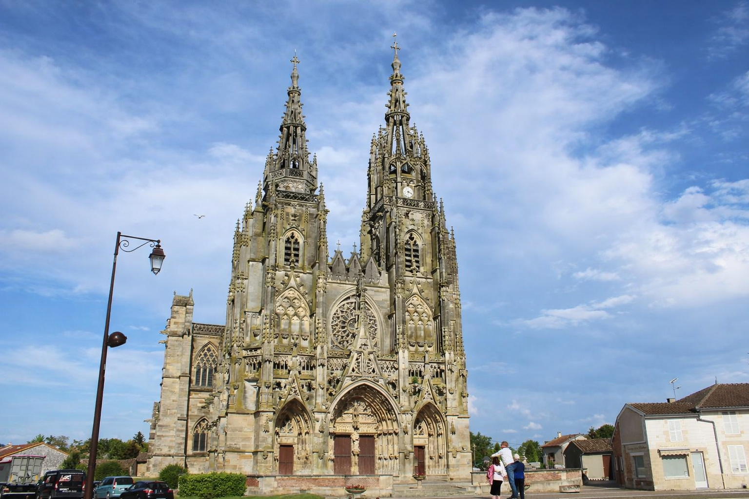 Gotische Basilika Notre-Dame de l'Épine mit reich verziertem Portal und zwei filigranen Türmen auf einem Dorfplatz, seitliches Straßenlaternenmotiv und Häuser im Hintergrund unter blauem Himmel mit leicht veränderten Wolken