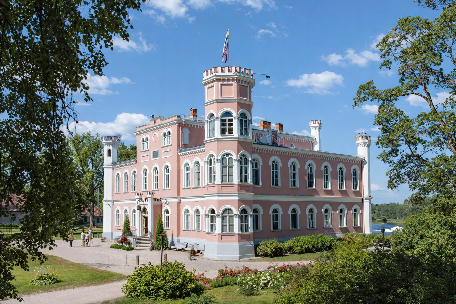 Rosa Schlossanlage Bīriņu pils mit weißen Fensterrahmen, Rundbogenfenstern und einem markanten Turm mit Fahne, umgeben von gepflegten Gärten, Blumenbeeten und altem Baumbestand, sonniges Licht mit klaren Schatten und leicht veränderte Wolken am blauen Himmel