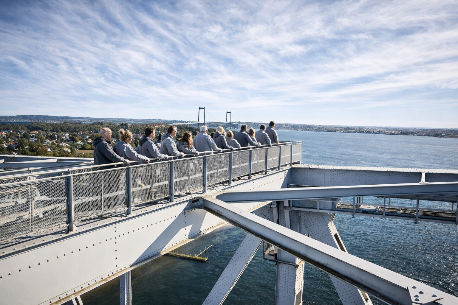 Teilnehmer beim Bridgewalking Lillebælt auf dem Stahlträger der Alten Lillebæltbrücke, gesichert entlang eines Geländers mit Blick über den Lillebælt und die Küstenlandschaft, im Hintergrund die Neue Lillebæltbrücke, darüber ein weiter Himmel mit feinen, nebligen Zirruswolken bei klarem Tageslicht