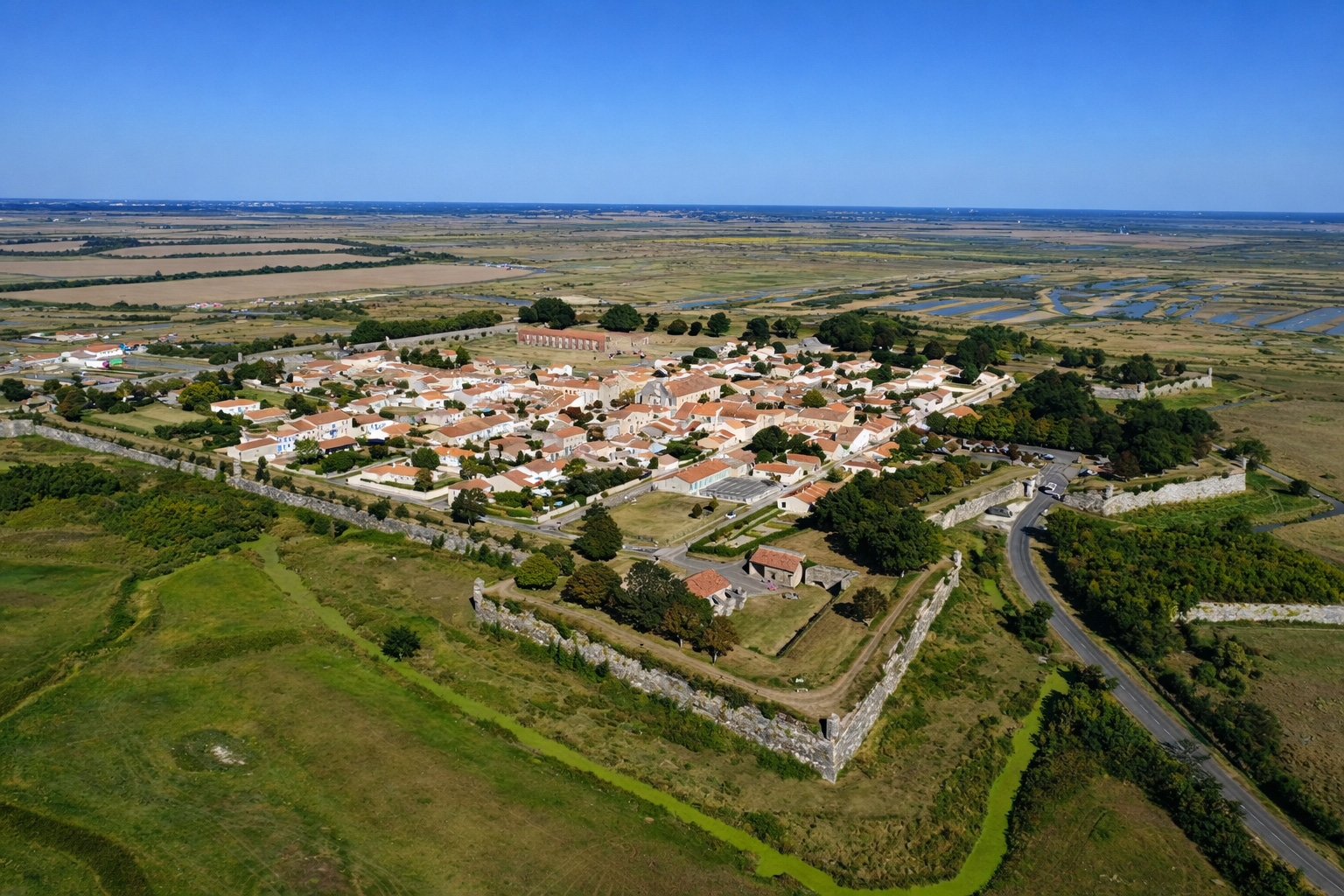 Luftaufnahme der befestigten Ortschaft Brouage mit sternförmigen Festungsmauern, roten Ziegeldächern und grünem Wassergraben, eingebettet in weite Felder und Salzwiesen unter klarem blauem Himmel
