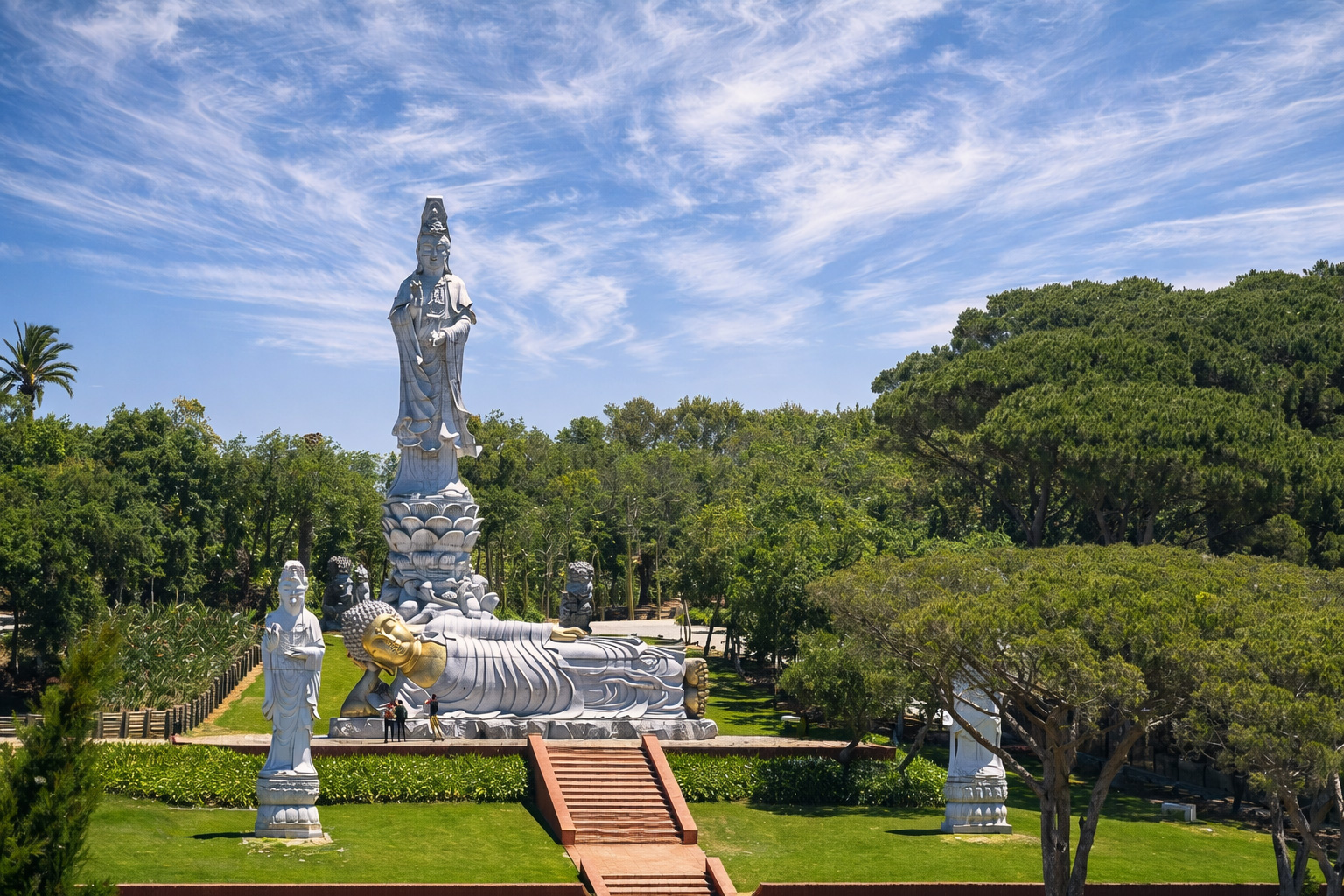 Blick auf den Buddha Eden Garden mit großer stehender Guanyin-Statue auf Lotus-Sockel und liegendem Buddha mit goldenem Kopf, davor breite rote Treppe und gepflegte Rasenflächen, umgeben von hohen Bäumen unter einem Himmel aus feinen, schleierartigen Zirruswolken