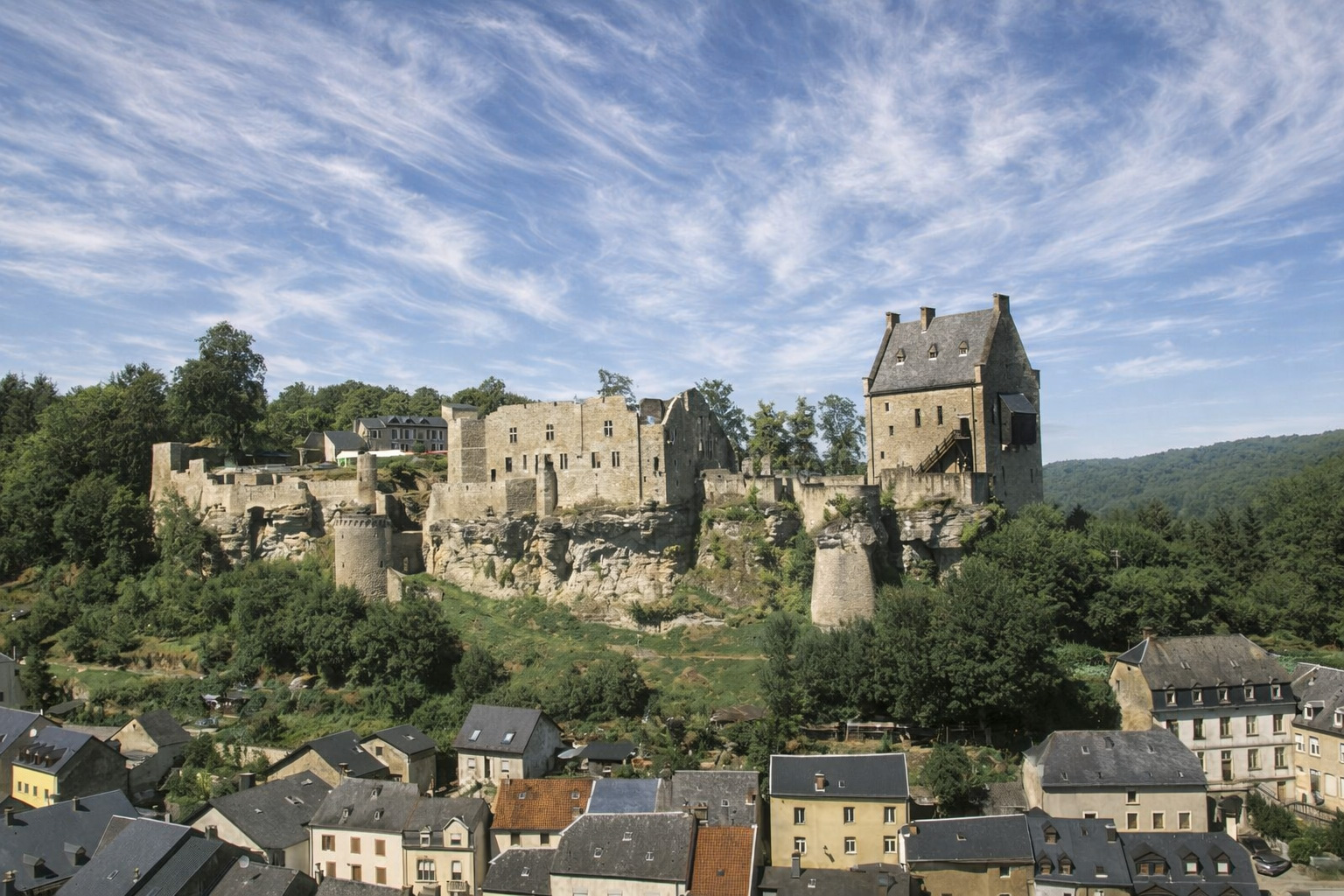 Burg Larochette auf einem felsigen Plateau über dem Ort Larochette in Luxemburg, mittelalterliche Ruinen und Wohnturm im hellen Sonnenlicht mit markanten Schatten, darunter dicht stehende Häuser und bewaldete Hügel unter einem von zarten Zirruswolken durchzogenen Himmel