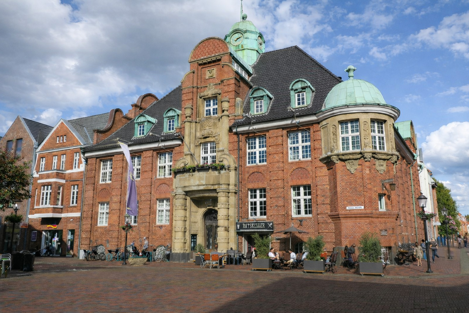 Historischer Marktplatz in Buxtehude mit eindrucksvollem Backstein-Rathaus, grünen Kupferkuppeln und reich verziertem Portal, Außengastronomie vor dem Ratskeller und Kopfsteinpflaster unter freundlichem Himmel