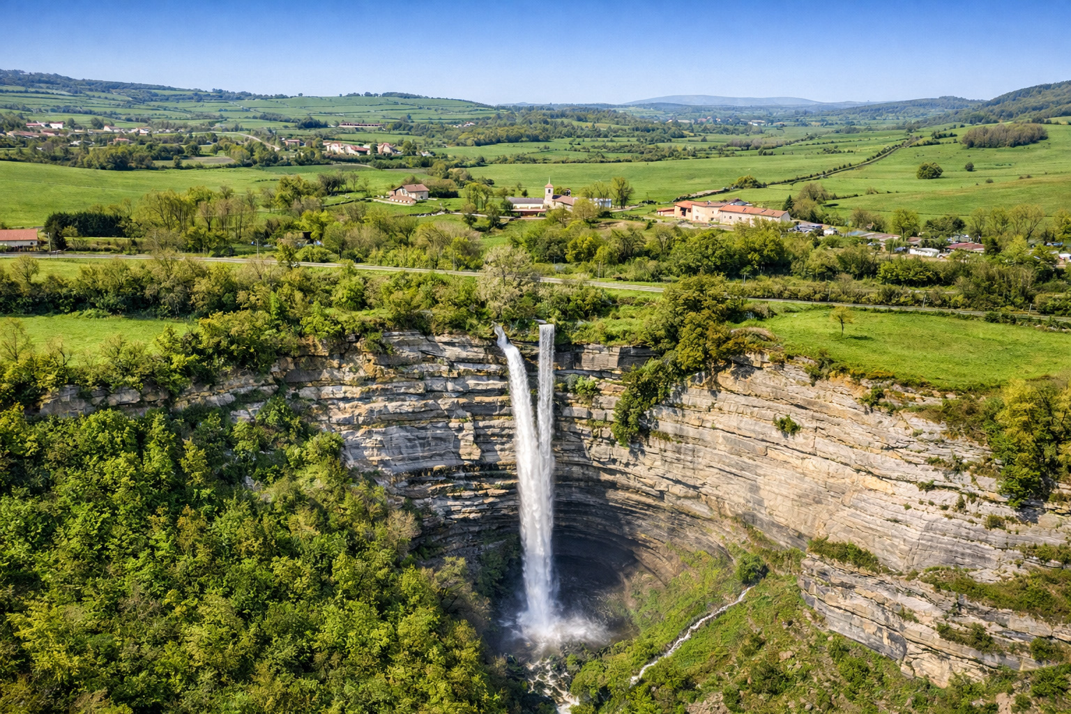 Luftaufnahme der Cascada de Gujuli, ein hoher Wasserfall stürzt in eine runde Felsmulde, umgeben von grünen Wäldern und Weiden; dahinter eine ländliche Hügellandschaft mit verstreuten Bauernhöfen unter klarem blauem Himmel