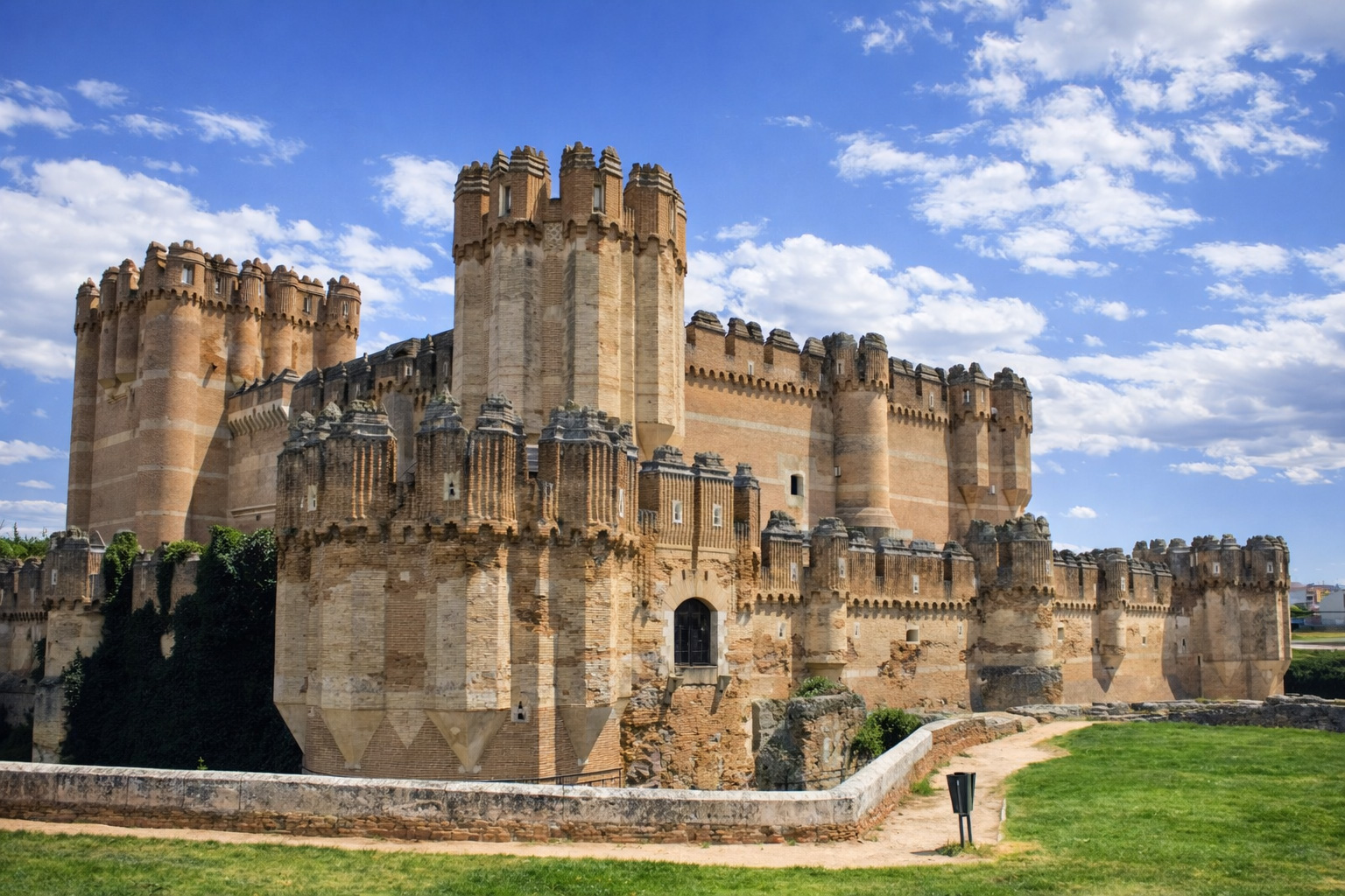 Castillo de Coca mit mächtigen Backsteinmauern, runden und eckigen Türmen sowie Zinnen und Wehrgängen, davor ein Weg am Burggraben und eine grüne Rasenfläche, darüber ein Himmel mit leicht veränderten, lockeren Wolken