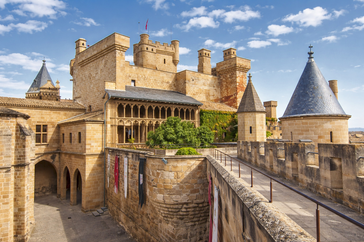 Innenhofansicht des Castillo de Olite mit goldfarbenen Sandsteinmauern, Zinnen und Türmen, einem Arkadengang mit filigranen Bögen und einer schmalen Wehrgang-Passage, dazu helles Sonnenlicht mit klaren Schatten und leicht aufgelockerte Wolken am Himmel