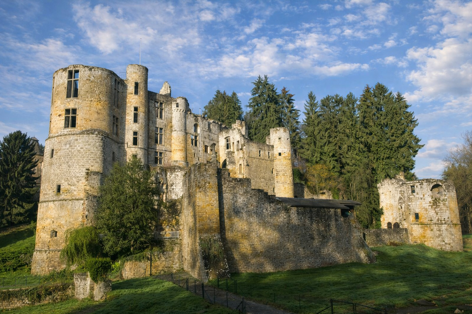 Château de Beaufort in Luxemburg mit massiven Rundtürmen und teils verfallenen Mauern, im Vordergrund eine grüne Wiese mit Weg und Zaun, dahinter hohe Nadelbäume, beleuchtet von klarem Sonnenlicht mit kontrastreichen Schatten unter leicht aufgelockerter Wolkendecke