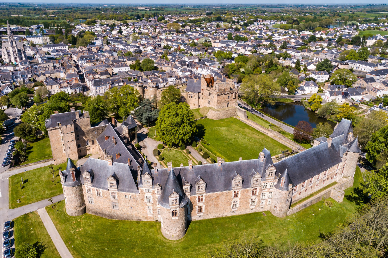 Luftaufnahme des Château de Châteaubriant mit langem Schlossflügel, runden Türmen und gepflegten Grünflächen, dahinter mittelalterliche Befestigungen und ein kleiner Teich am Rand der Altstadt unter kräftigem Sonnenlicht mit klaren Schatten