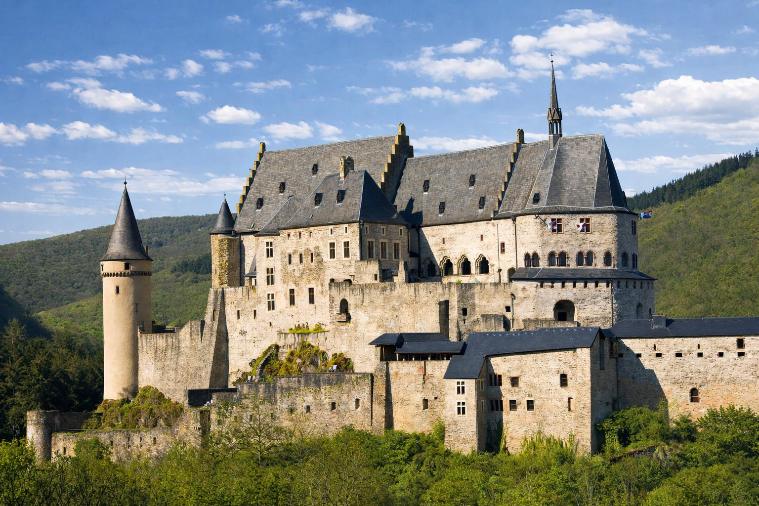 Château de Vianden hoch über dem Tal in Luxemburg, mächtige Burganlage mit hellen Steinmauern und dunklen Schieferdächern vor bewaldeten Hügeln, im Vordergrund frisches Grün, klare Sonne mit kräftigen Schatten unter einem blauen Himmel mit leicht veränderten, locker verteilten Wolken
