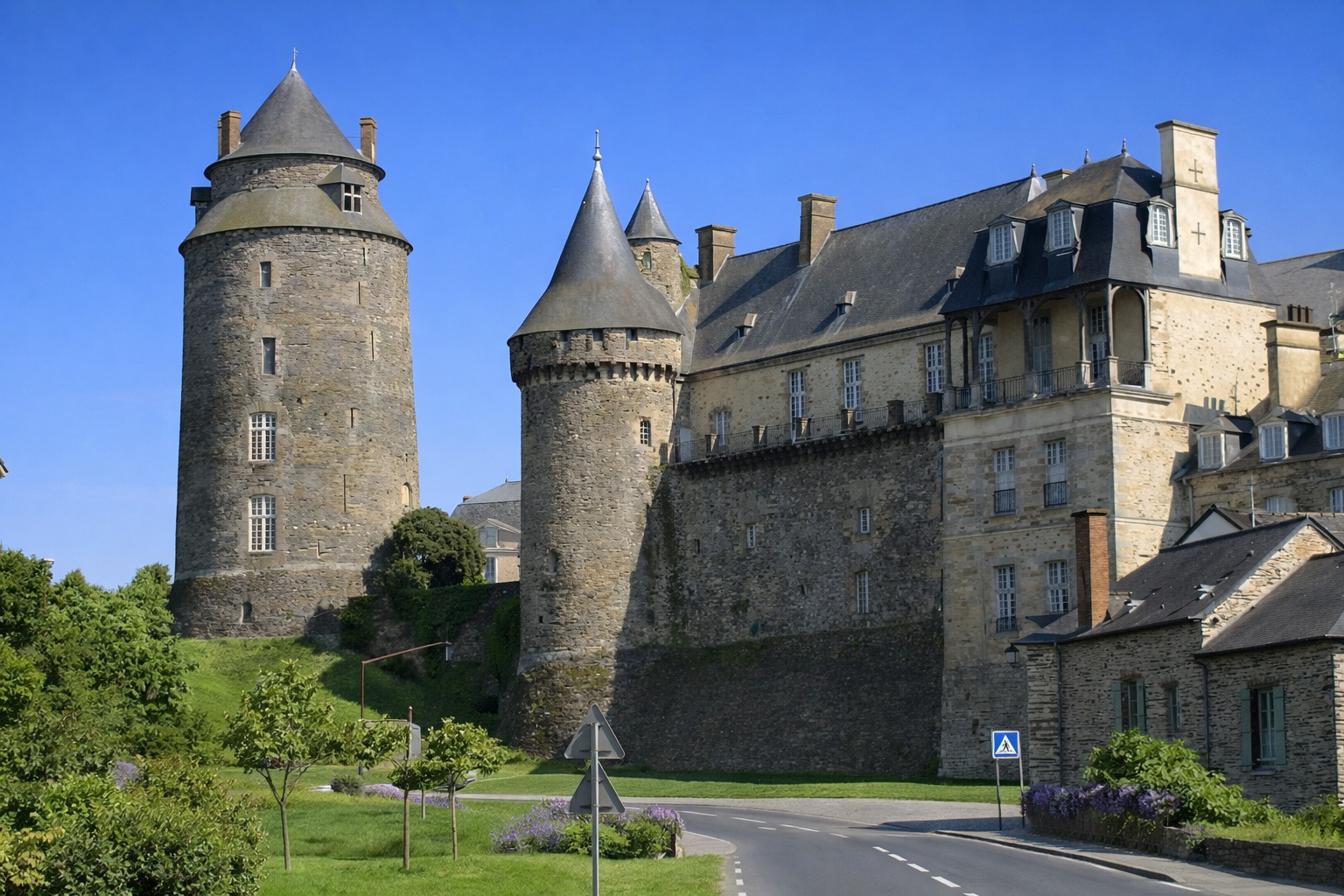 Mittelalterliches Schloss von Châteaugiron mit zwei runden Steintürmen und großem Herrenhaus mit Schieferdach, davor eine geschwungene Straße ohne Fahrzeuge und eine sattgrüne Wiese unter klarem blauem Himmel