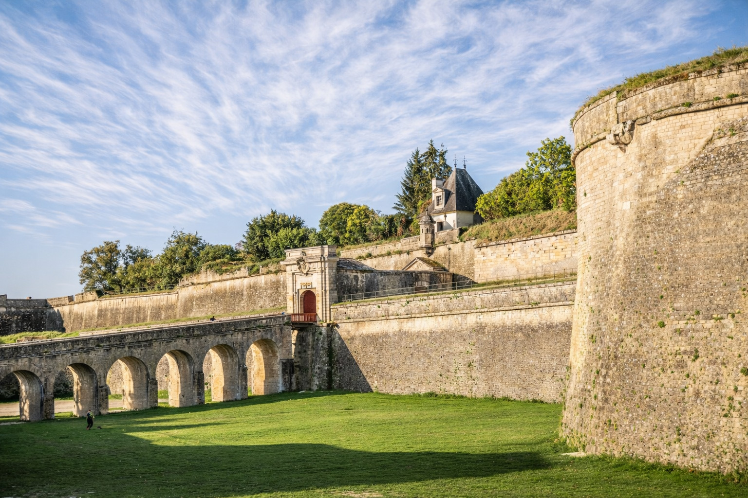 Massive Festungsmauern der Citadelle de Blaye mit steinerner Bogenbrücke und Toranlage über einer grünen Wiese, flankiert von Bäumen und einer kleinen Kapelle auf der Bastion unter einer großflächigen, neblig wirkenden Zirruswolke