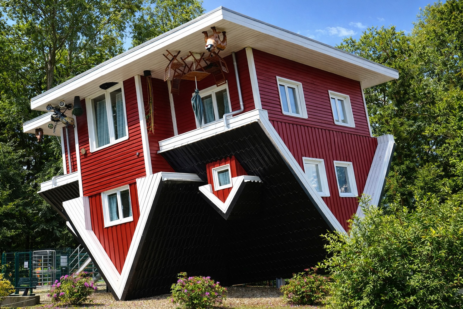 Schräg von vorn aufgenommenes, auf dem Kopf stehendes rotes Haus mit weißer Holzverkleidung im Tierpark Gettorf, das Dach liegt am Boden und Details wie Möbel und Dekoration hängen kopfüber unter dem Dachüberstand, umgeben von grünen Bäumen und Sträuchern bei klarem Sonnenlicht mit kräftigen Schatten