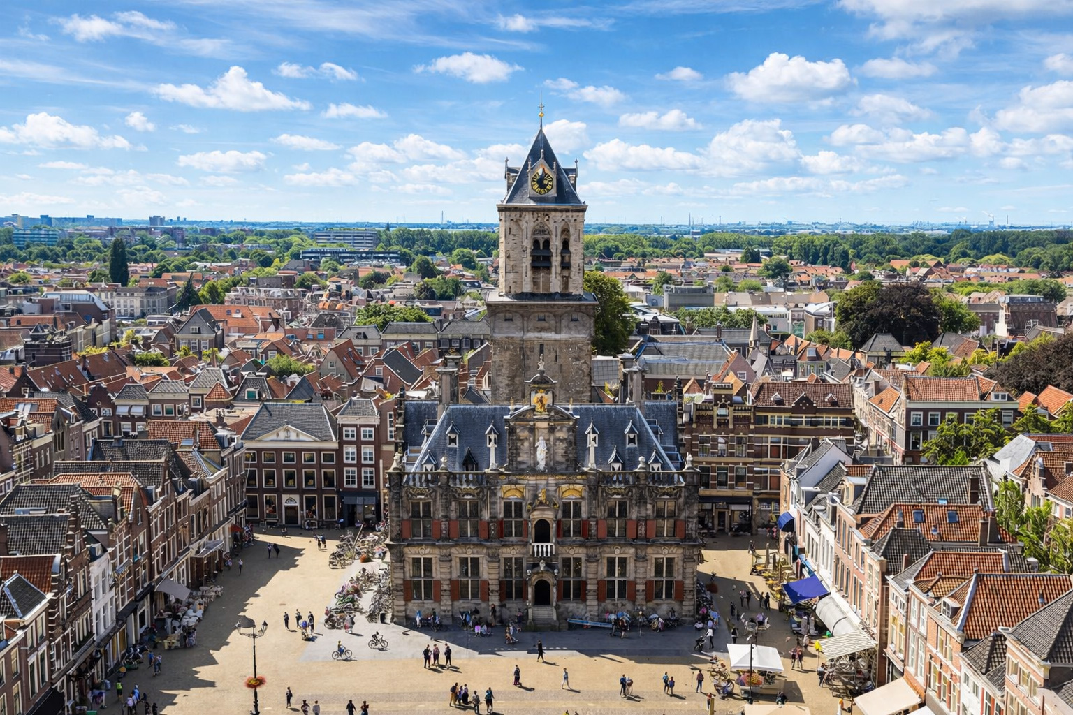 Luftblick auf den Marktplatz von Delft mit dem historischen Rathaus und seinem hohen Uhrturm, umgeben von dicht stehenden Giebelhäusern und roten Ziegeldächern, belebt von Fußgängern und Radfahrern bei klarem Sonnenlicht und leicht veränderten Wolken am Himmel