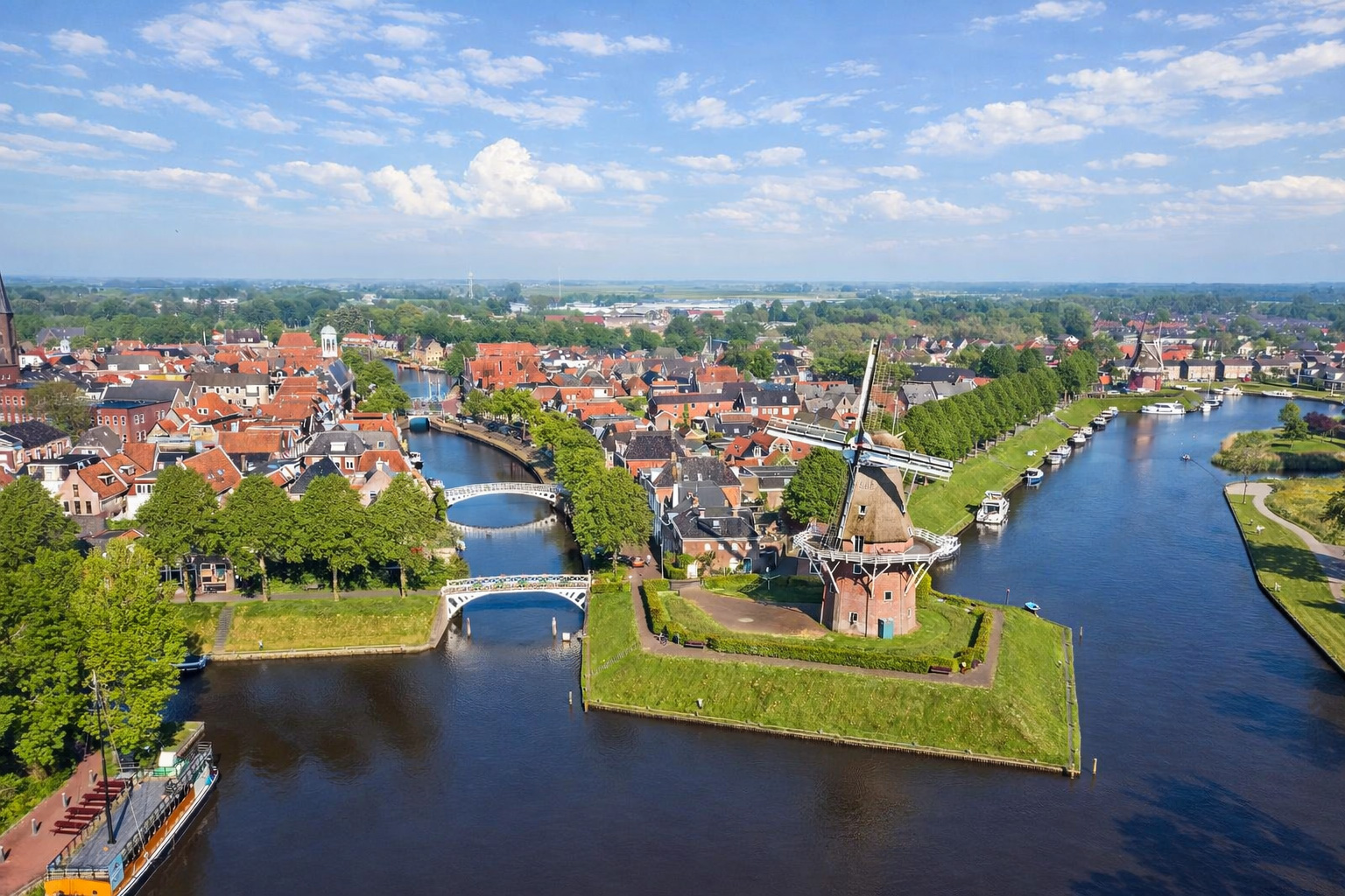 Luftaufnahme von Dokkum mit roten Ziegeldächern, geschwungenen Kanälen und mehreren Brücken, im Vordergrund eine historische Windmühle auf einer grünen Landzunge, Boote liegen entlang der Uferwege, darüber ein heller Himmel mit leicht veränderten Wolken im klaren Sonnenlicht