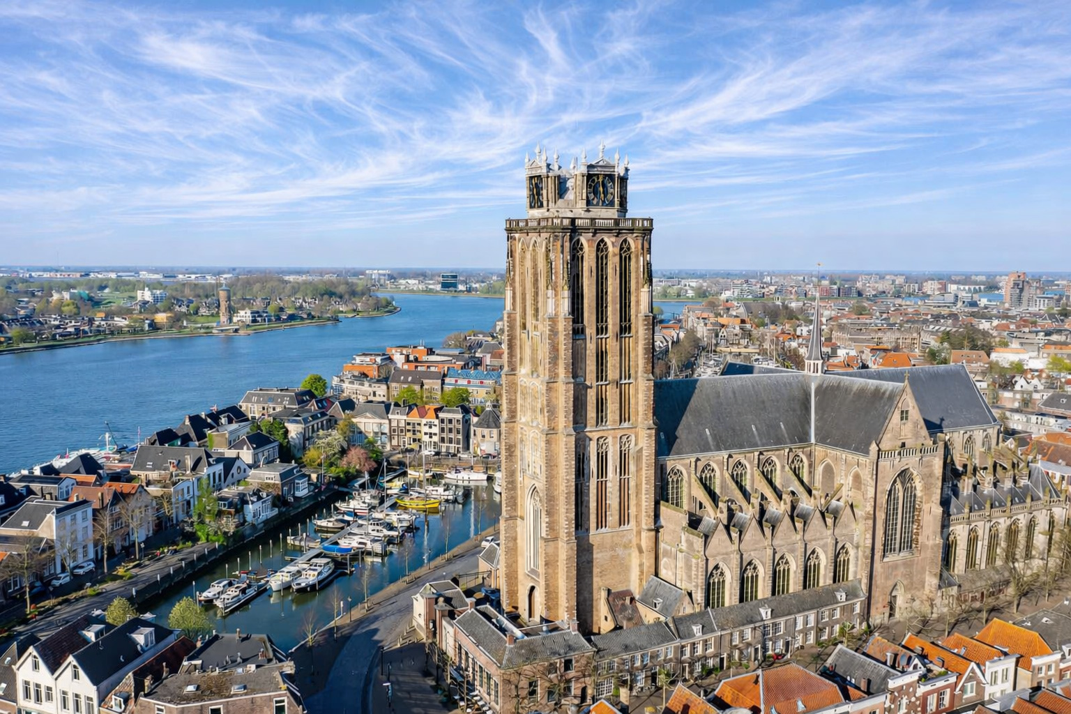 Luftaufnahme von Dordrecht mit der markanten Grote Kerk und ihrem hohen Turm über den Giebelhäusern, Kanälen und Bootsanlegern am breiten Fluss, bei klarem Sonnenlicht und einem Himmel voller feiner, nebliger Zirruswolken