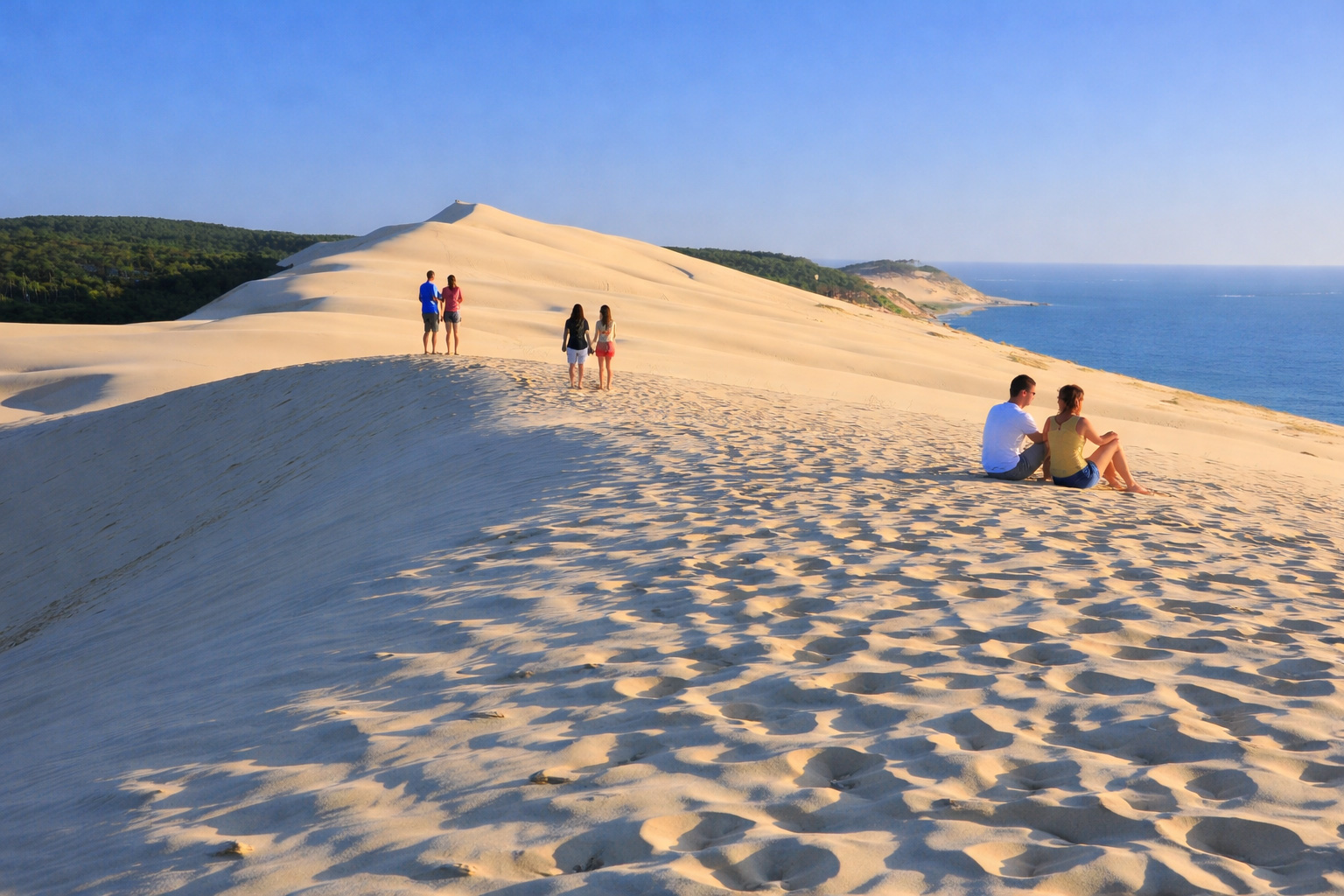 Weitläufiger Kamm der Dune du Pilat mit feinen Sandrippeln und tiefen Schatten, Spaziergänger und Sitzende auf der Dünenkante, dahinter Pinienwald und Blick auf den Atlantik unter klarem blauem Himmel