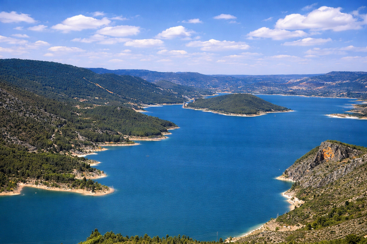 Panoramablick über den Embalse de Entrepeñas mit tiefblauem Stausee, geschwungenen Uferlinien und bewaldeten Hügeln, im Hintergrund weite Berglandschaft unter leicht veränderten, lockeren Wolken