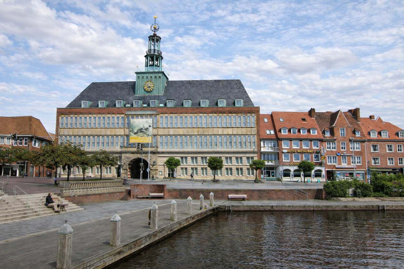 Blick auf das historische Rathaus am Wasser in Emden mit markantem Uhrturm, roten Backsteinhäusern und Uferpromenade, spiegelnde Kanalfläche im Vordergrund.