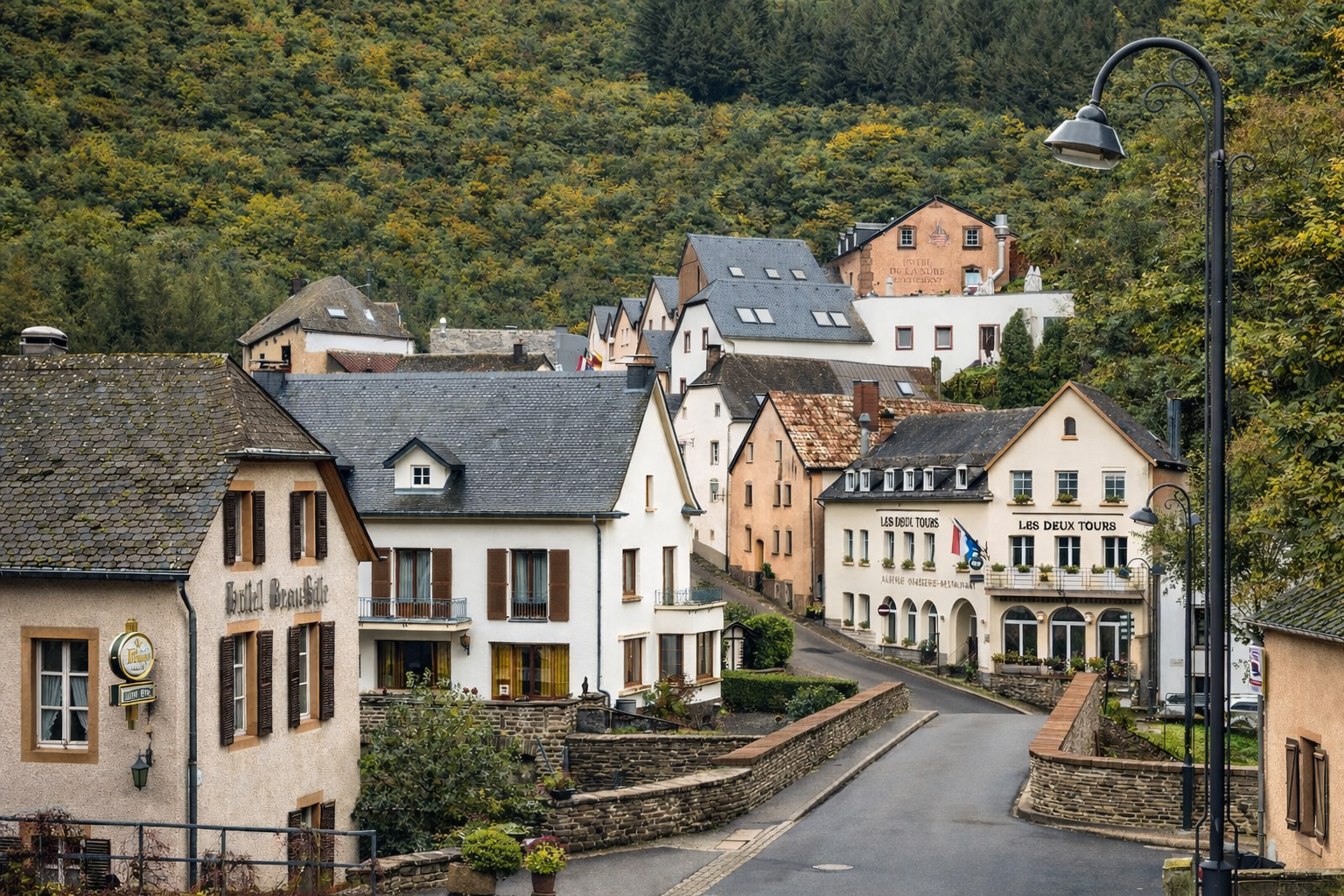 Blick auf Esch-sur-Sûre in Luxemburg mit gepflegten Häusern und Hotels entlang einer schmalen Straße über eine kleine Brücke, eingerahmt von dicht bewaldeten Hängen in herbstlichen Farben, klares Sonnenlicht mit kräftigen Schatten und ruhige, menschenleere Szenerie