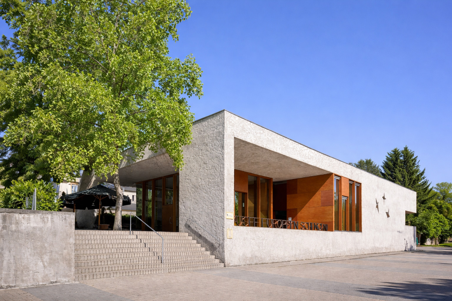 Moderne, kubische Architektur des Europäischen Museums in Schengen mit heller Betonfassade, großen Holz- und Glasflächen und breiter Treppe im Vordergrund, eingerahmt von Bäumen unter klarem blauem Himmel