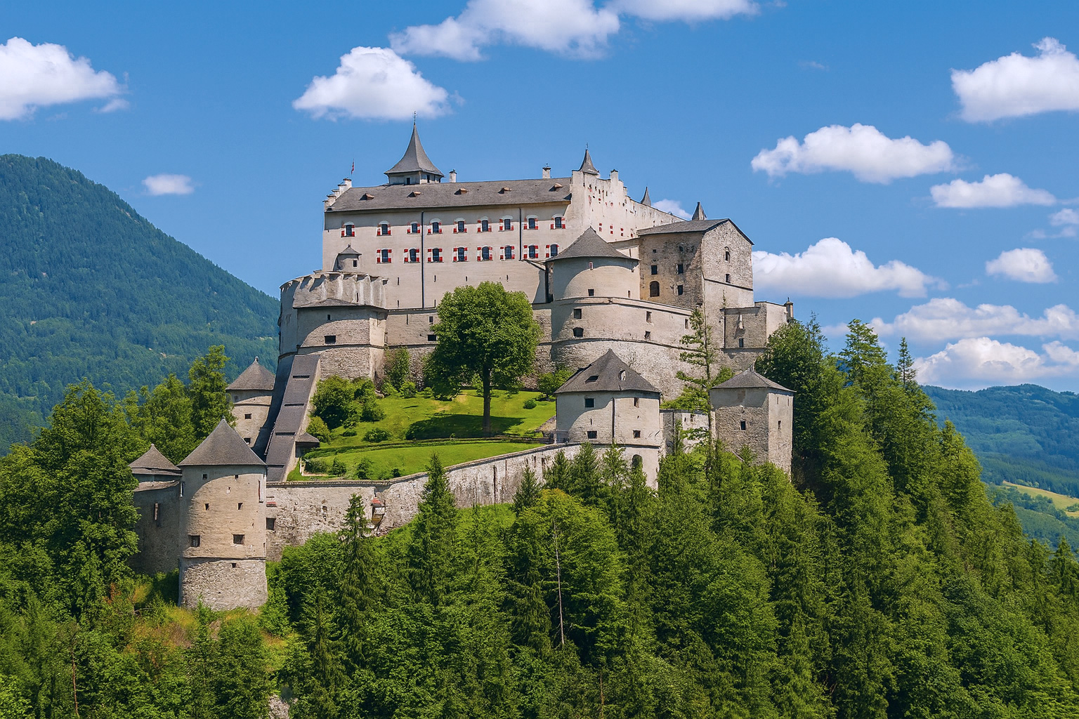 Festung Hohenwerfen auf einem bewaldeten Hügel bei klarem Wetter mit blauem Himmel und weiß-grauen Wolken im besten Sonnenlicht