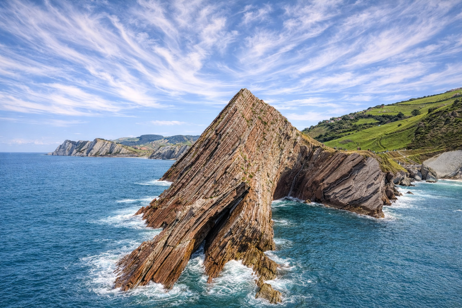Schräg aufragende Flysch-Felsformationen an der Küste von Zumaia, von Wellen umspült und von grünen Hängen begleitet, mit weitem Blick über das tiefblaue Meer unter einer großflächigen, neblig wirkenden Zirruswolke