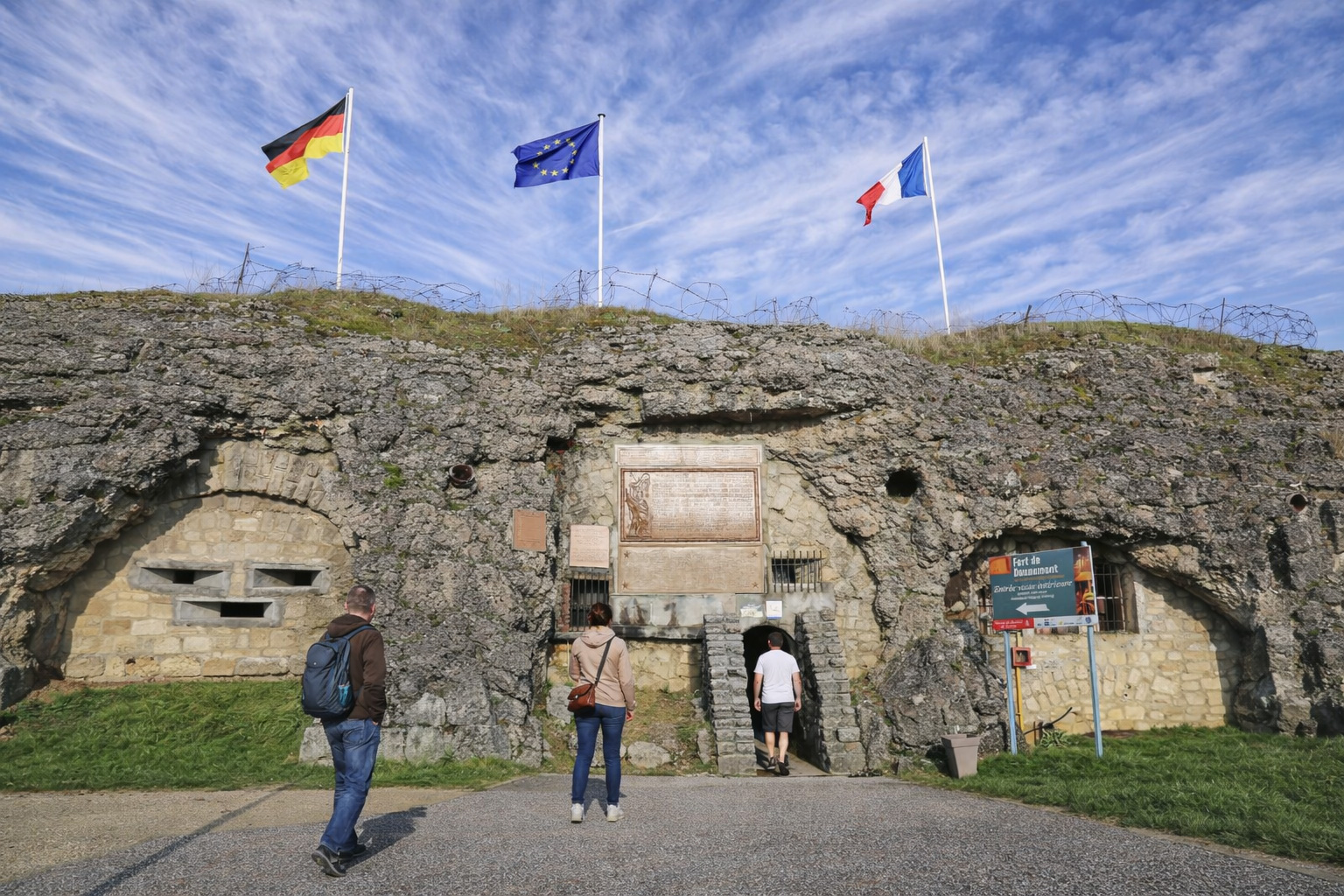 Eingangsfront des Fort de Douaumont mit in Fels und Beton eingelassenen Öffnungen, Gedenktafeln und Informationstafel, darüber Flaggen von Deutschland, der Europäischen Union und Frankreich sowie Stacheldraht unter einem hohen Himmel mit feinen Schleierwolken