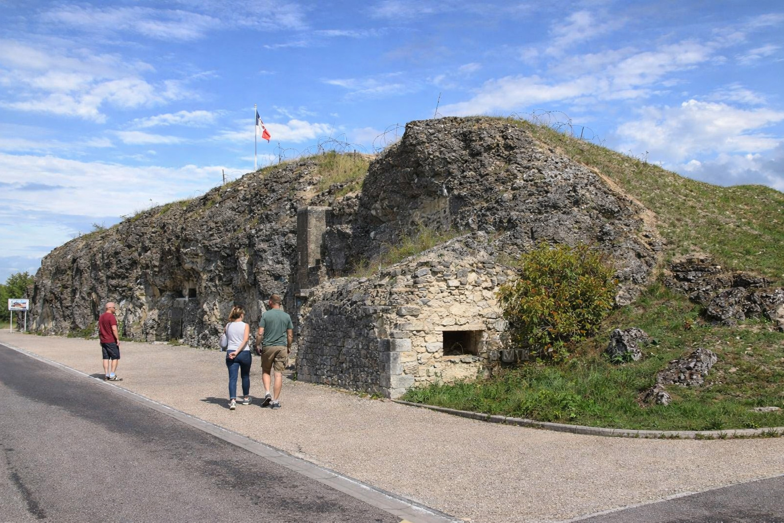 Blick auf das Fort de Vaux mit felsiger, grasbewachsener Befestigung und kleinem gemauerten Vorbau am Weg, französische Flagge auf dem Hügel und Stacheldraht auf der Kuppe unter blauem Himmel mit leicht veränderten Wolken