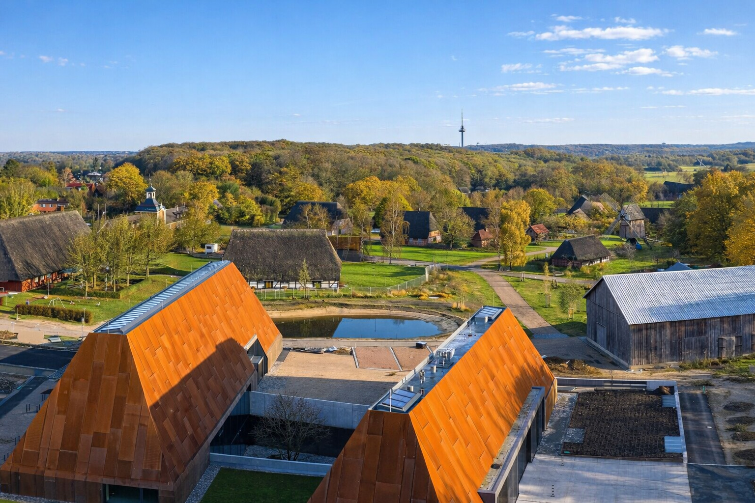 Luftaufnahme des Freilichtmuseums Molfsee bei Kiel mit modernen kupferfarbenen Museumsgebäuden im Vordergrund, einem Teich und Wegen dazwischen sowie historischen Reetdachhäusern und einer Windmühle in der grünen Kulturlandschaft unter blauem Himmel mit lockeren Wolken