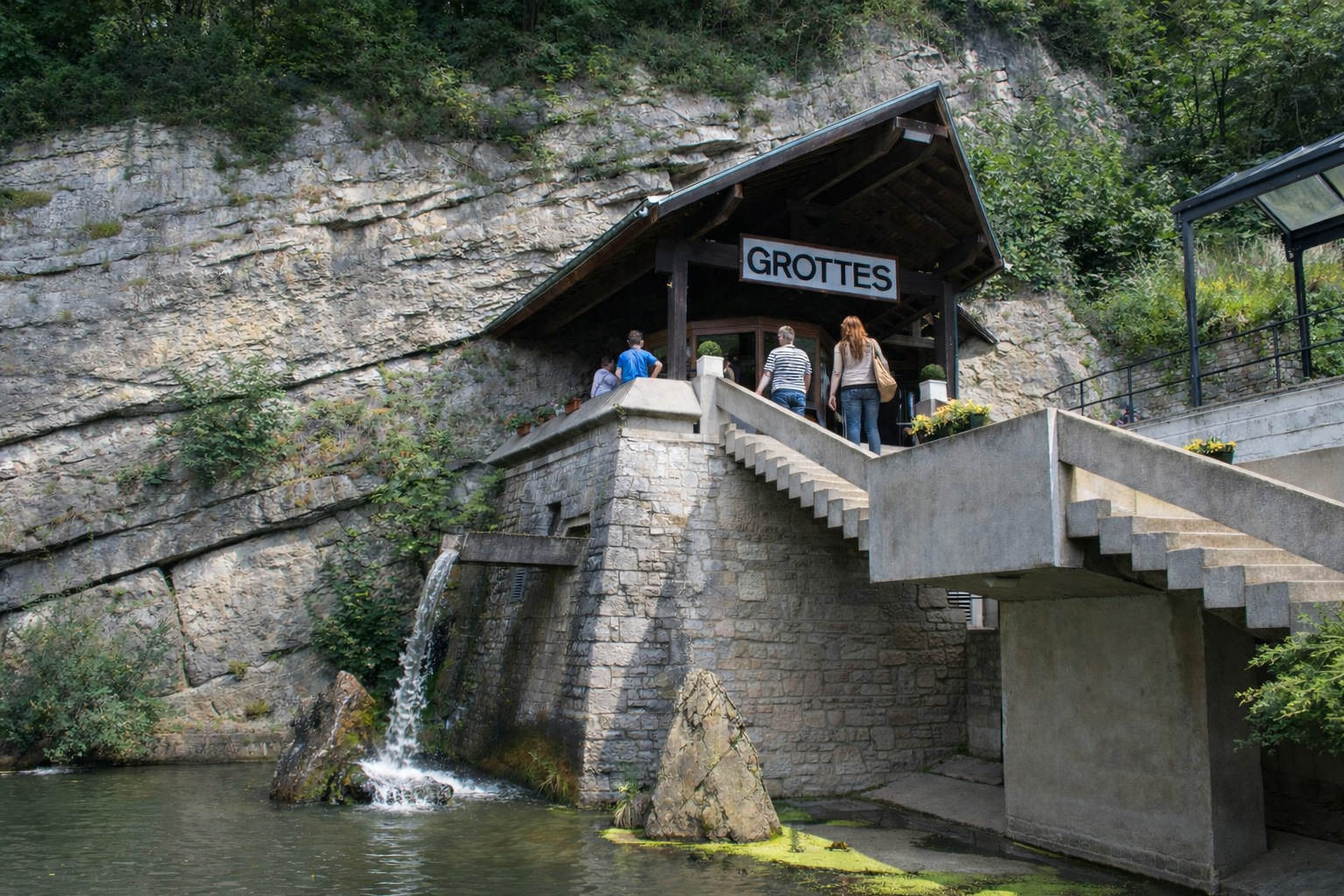 Eingang der Grottes de Remouchamps an einer steilen Felswand mit Holzüberdachung und Treppenanlage, darunter ein kleiner Wasserfall, der in ein Becken fließt, umgeben von dichtem Grün, klares Sonnenlicht mit kontrastreichen Schatten und Besuchergruppe am Zugang