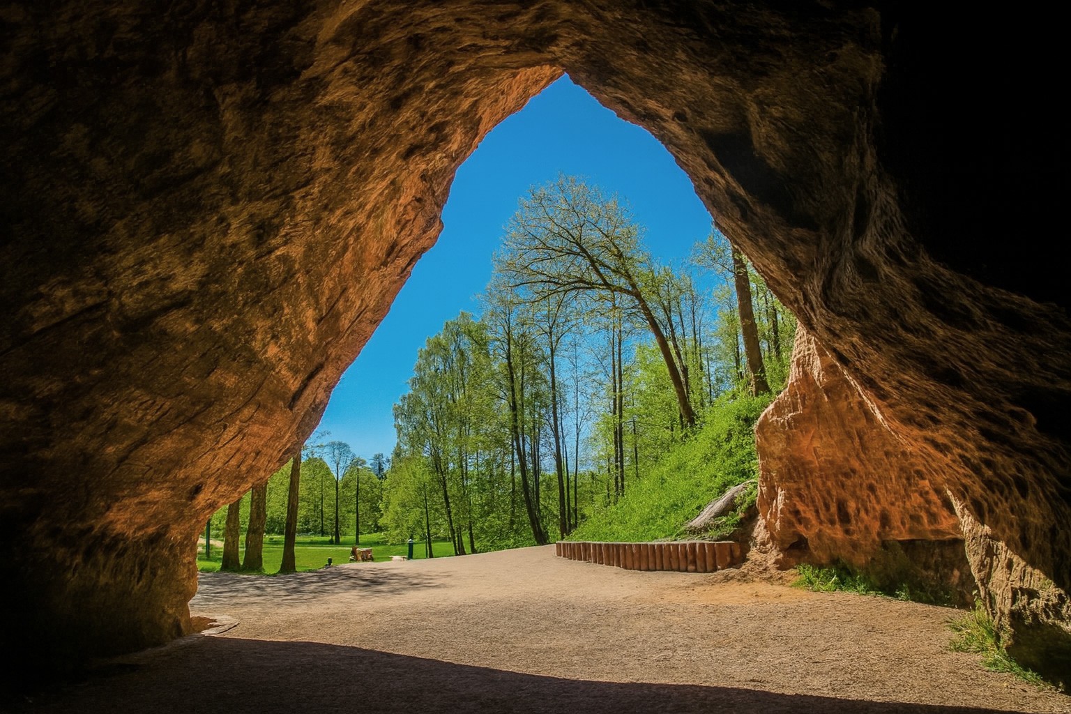 Blick aus der Gutman’s Höhle bei Sigulda auf den sonnigen Park mit grünen Bäumen unter einem wolkenlosen blauen Himmel.