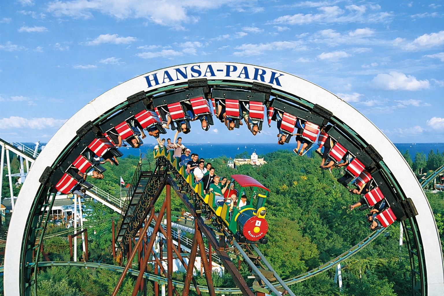 Achterbahn im Hansa-Park mit großem Looping und Schriftzug über der Strecke, Zug mit rot-weiß gestreiften Sitzen in luftiger Höhe, Blick über grüne Baumkronen bis zur Ostsee am Horizont unter blauem Himmel