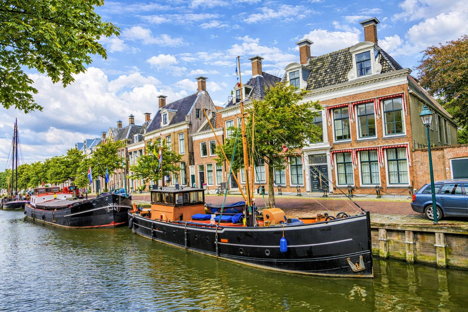 Idyllische Kanalszene in Harlingen mit festgemachten historischen Booten im Vordergrund, ruhigem Wasser mit Spiegelungen und einer Reihe traditioneller Häuser mit Giebeln und Ziegeldächern entlang der Uferpromenade, aufgenommen bei klarem Sonnenlicht unter einem Himmel mit leicht veränderten Wolken