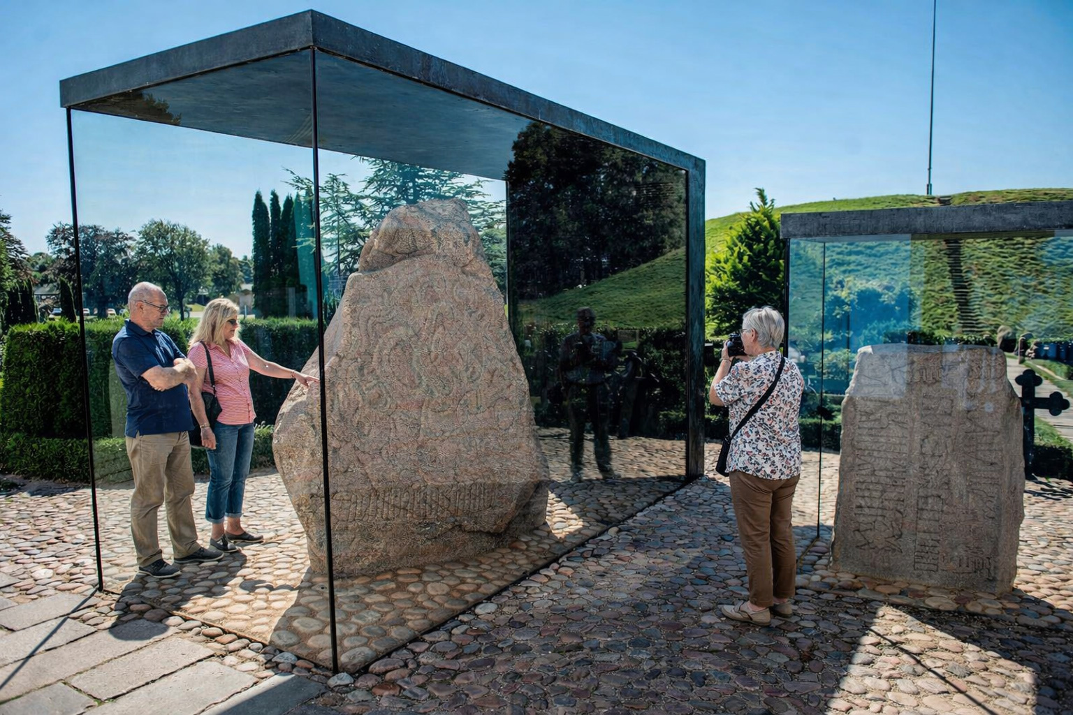 Runensteine der Jelling Monumente in modernen Glasvitrinen auf einem gepflasterten Platz, der große Stein steht zentral im transparenten Kubus, rechts ein weiterer Runenstein in einer kleineren Glashülle, Besucher betrachten und fotografieren die Steine, im Hintergrund gepflegte Grünanlagen und ein grasbewachsener Hügel unter klarem Himmel